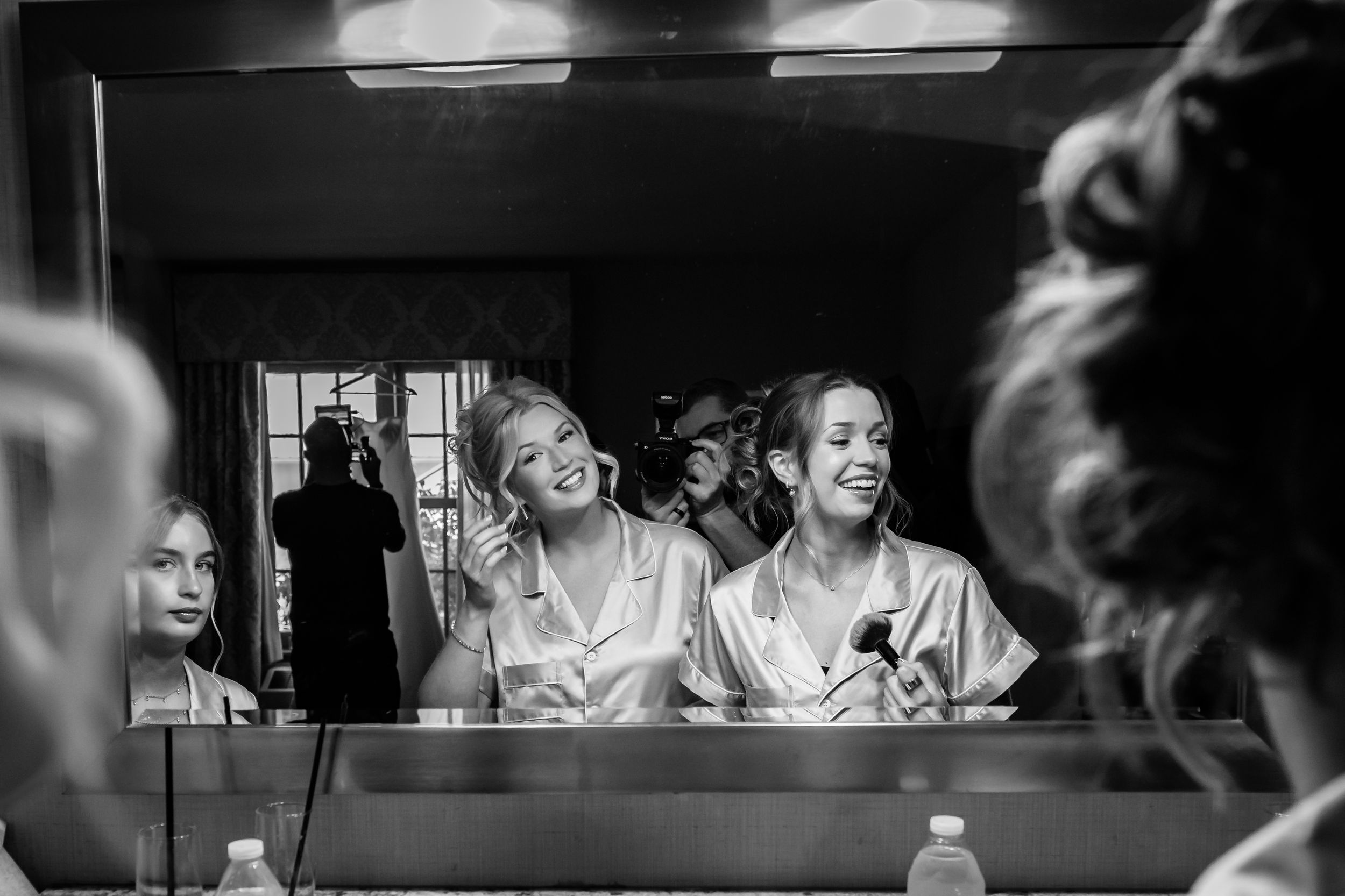 Bridesmaids laughing and applying makeup together reflected in mirror during wedding prep.