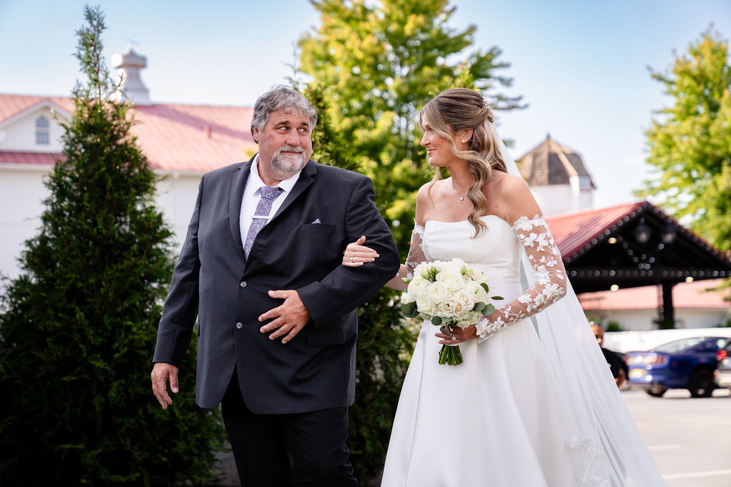 Father escorts smiling bride in lace wedding gown carrying white bouquet to ceremony.