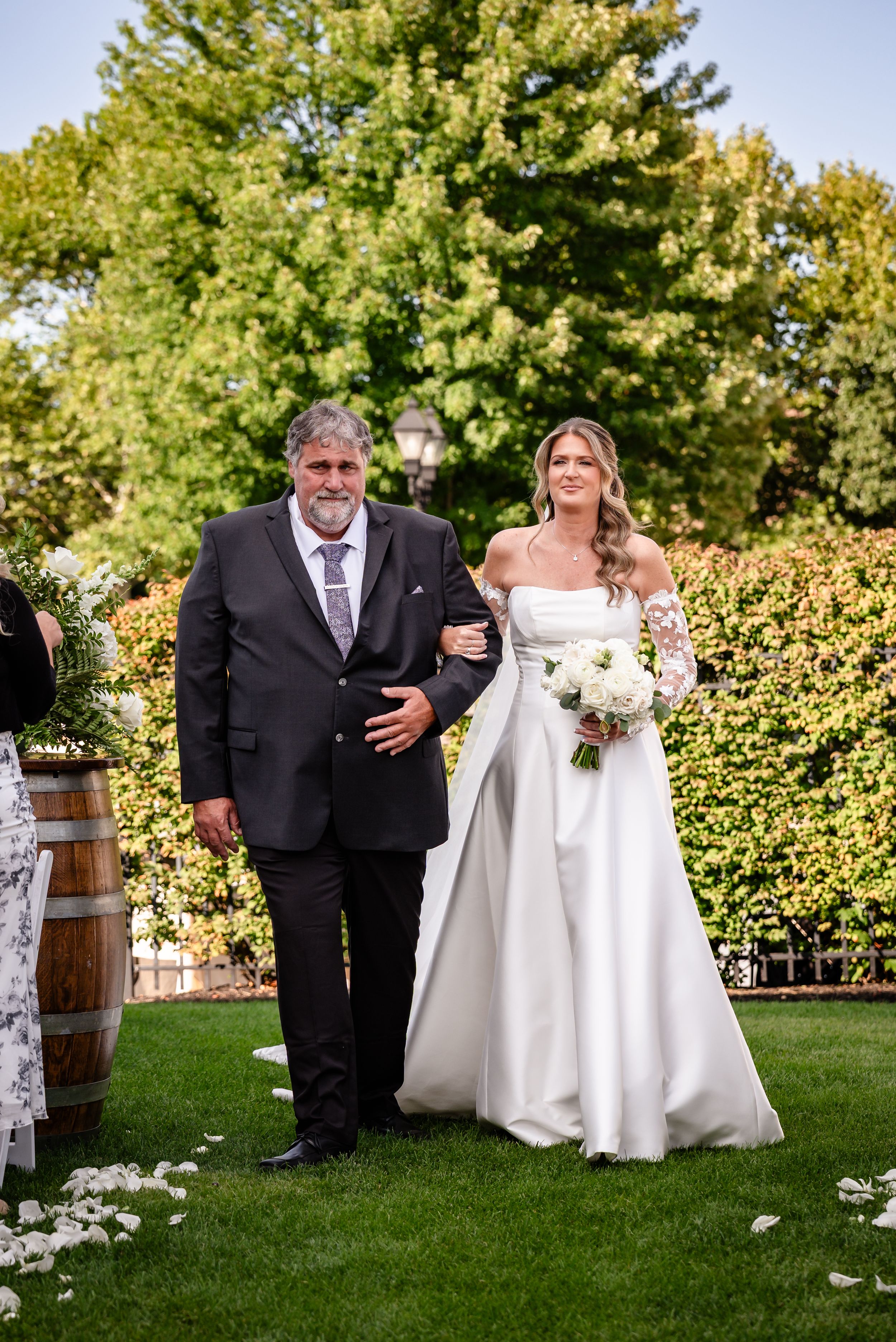 Bride in white gown walks down outdoor aisle with escort holding bouquet at garden wedding ceremony.