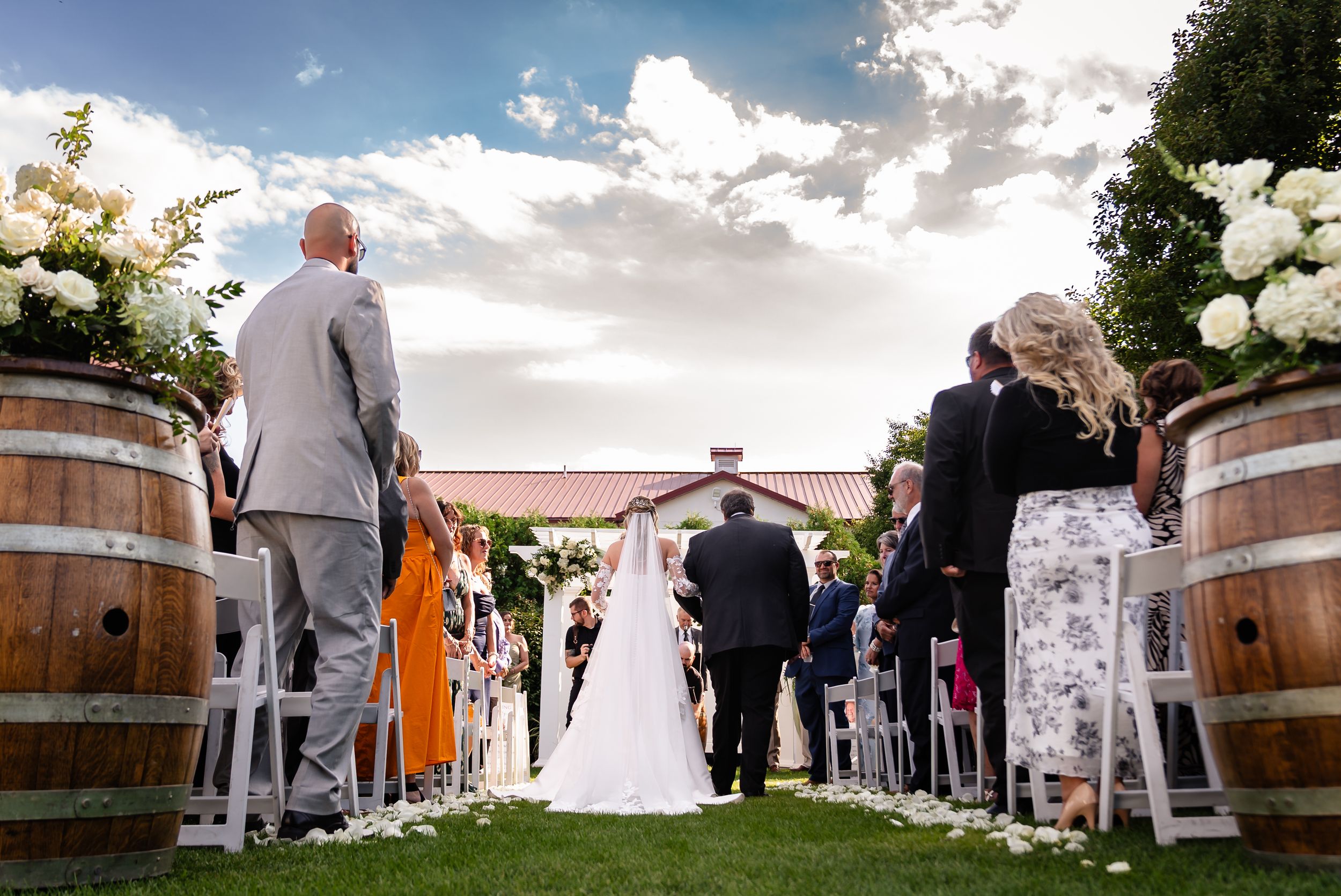 Bride walks down outdoor aisle flanked by wine barrels at vineyard wedding ceremony.