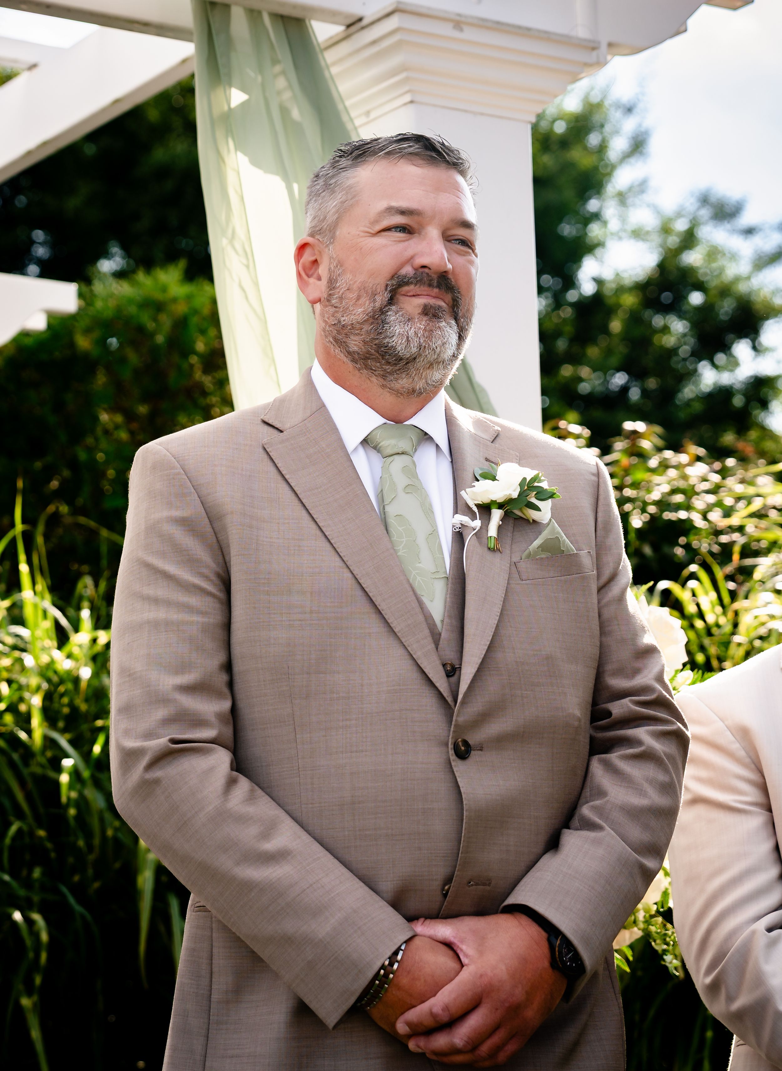 Groom in tan suit with green tie and boutonniere smiling outdoors at wedding ceremony.