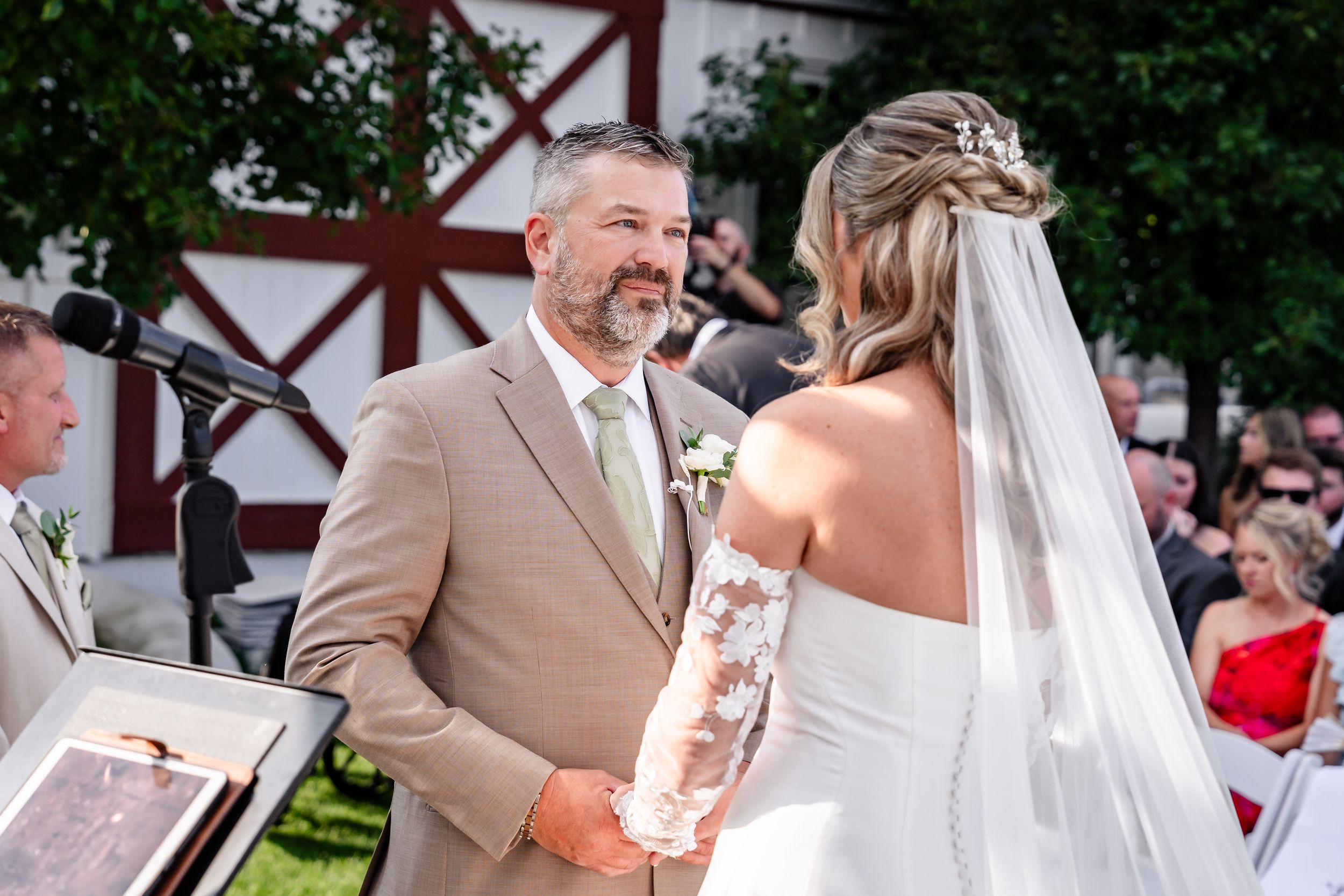 Groom in tan suit smiles lovingly at bride during outdoor wedding ceremony near a rustic red barn.