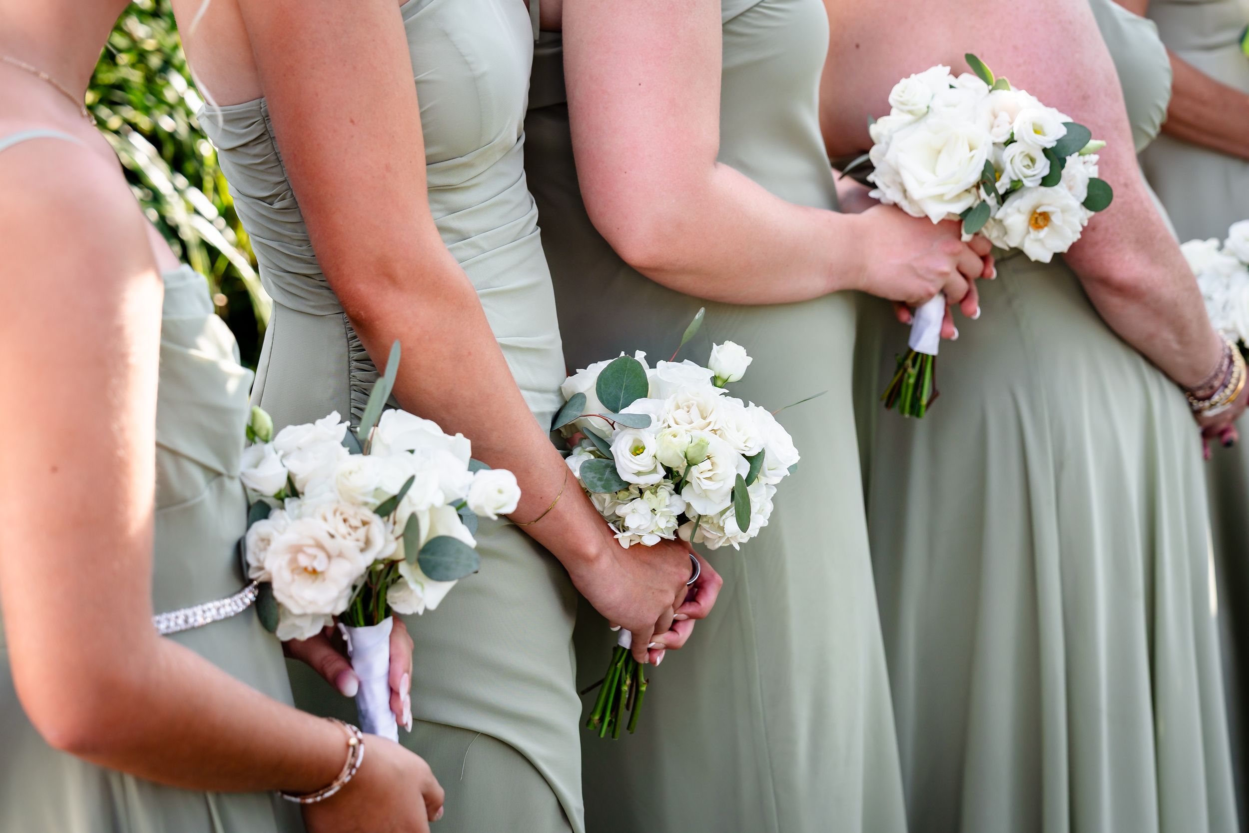 Bridesmaids in sage green dresses holding elegant white rose and eucalyptus bouquets at outdoor wedding.
