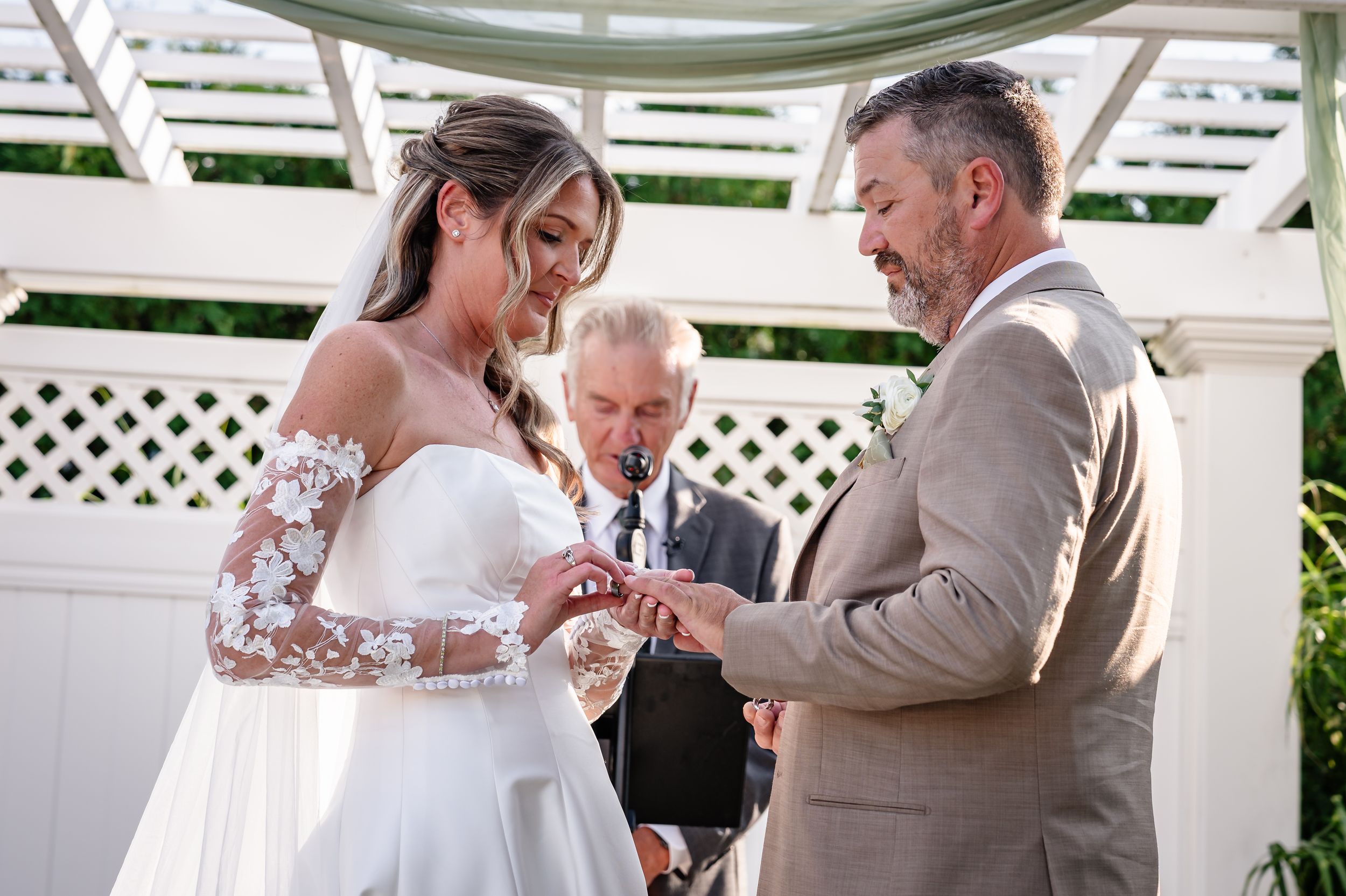 Groom places wedding ring on brides finger during outdoor ceremony beneath a white pergola.