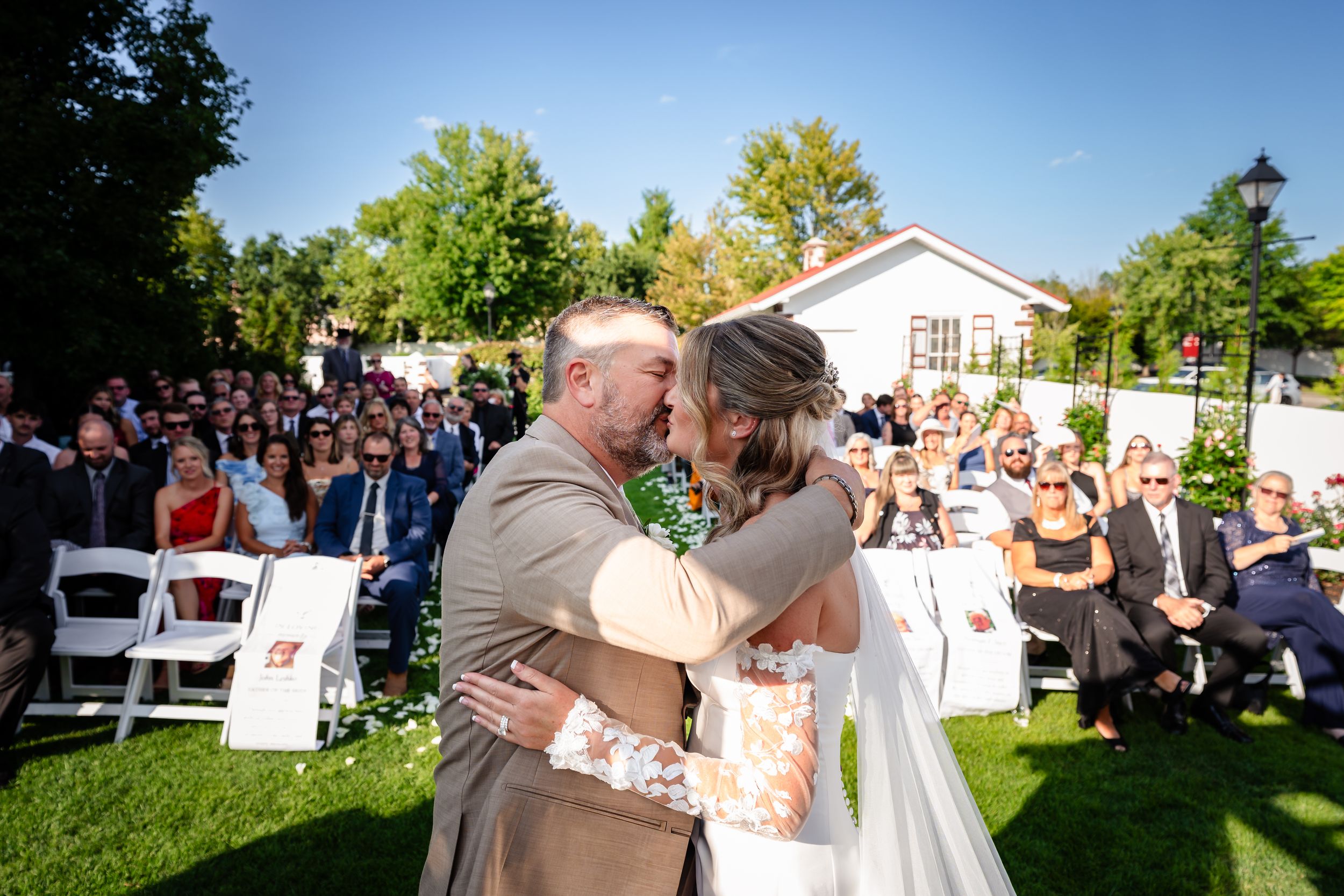 Groom embraces bride at outdoor wedding ceremony with guests seated on white chairs in background.