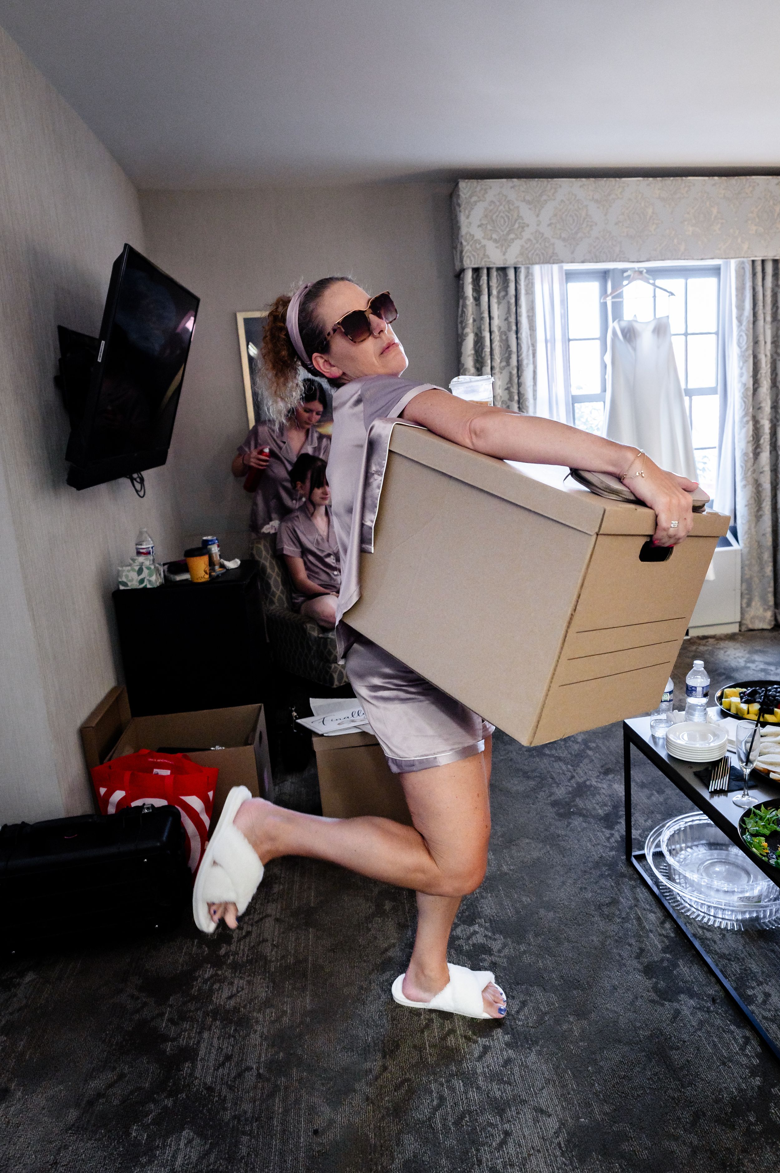 Bridesmaid posing playfully carrying a cardboard box in hotel room with wedding dress hanging in background.