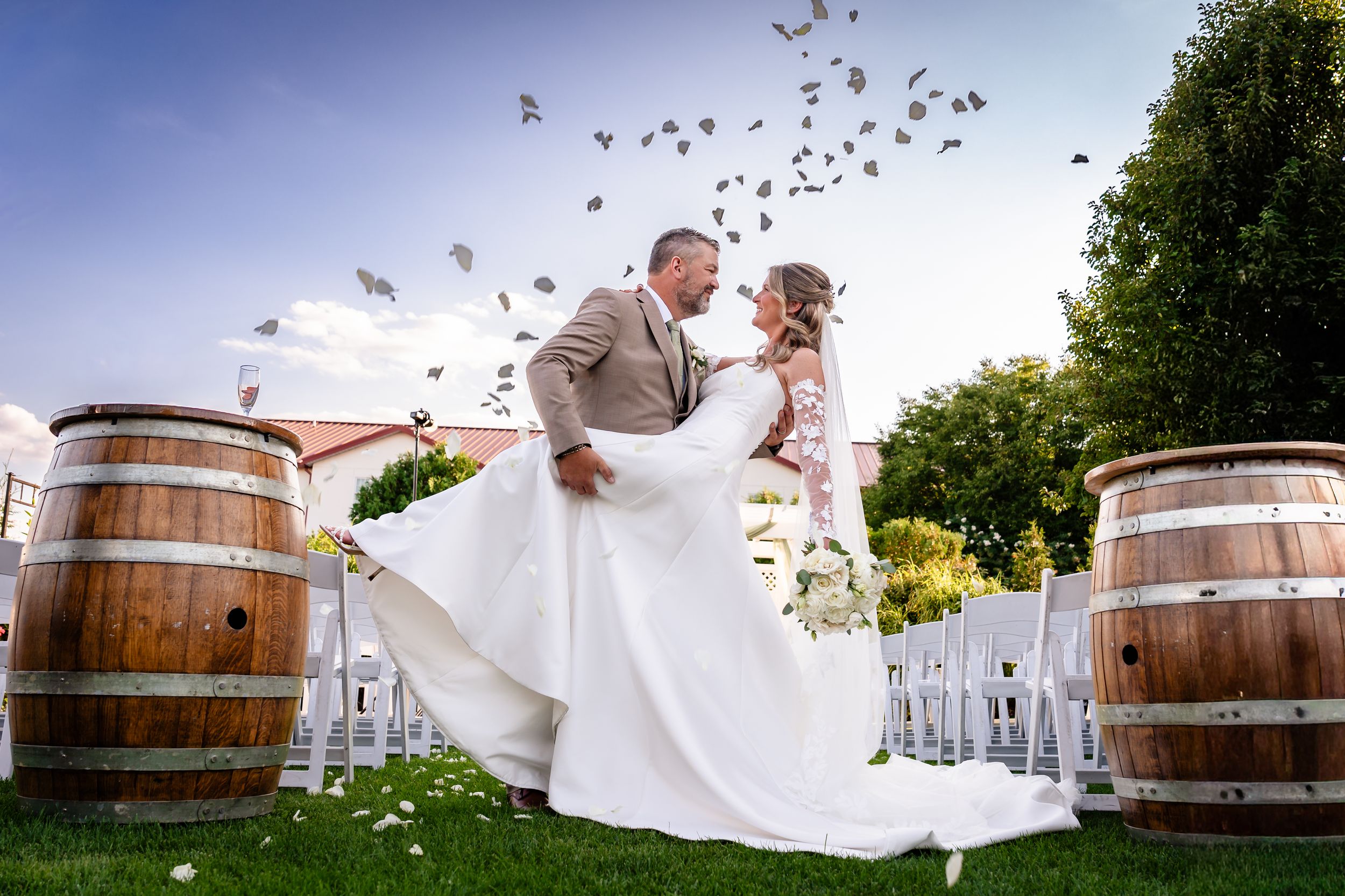 Groom lifts bride in white gown outdoors at winery wedding with wine barrels and flower petals falling.