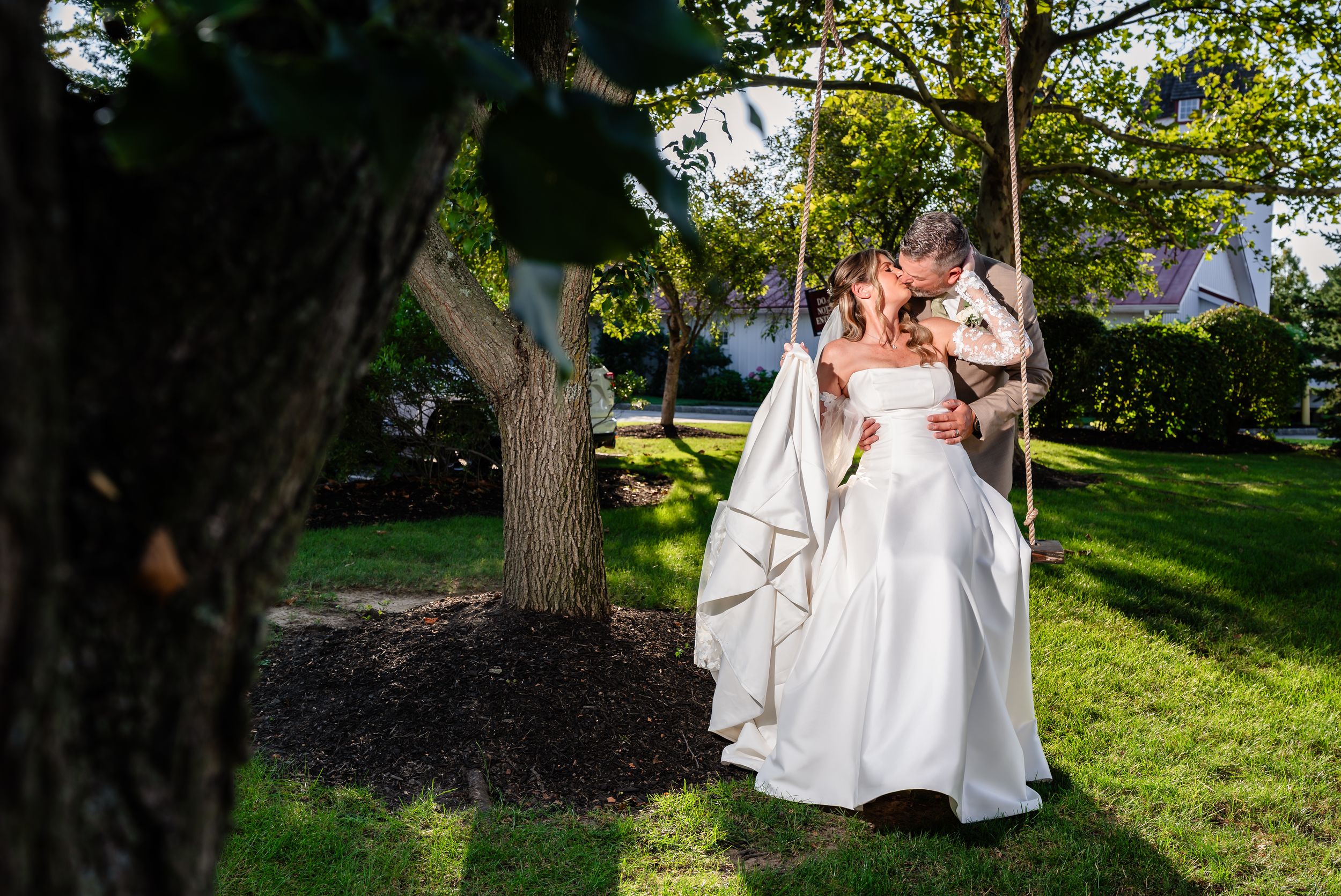 Bride and groom share a romantic kiss on a tree swing in a lush green garden on their wedding day.