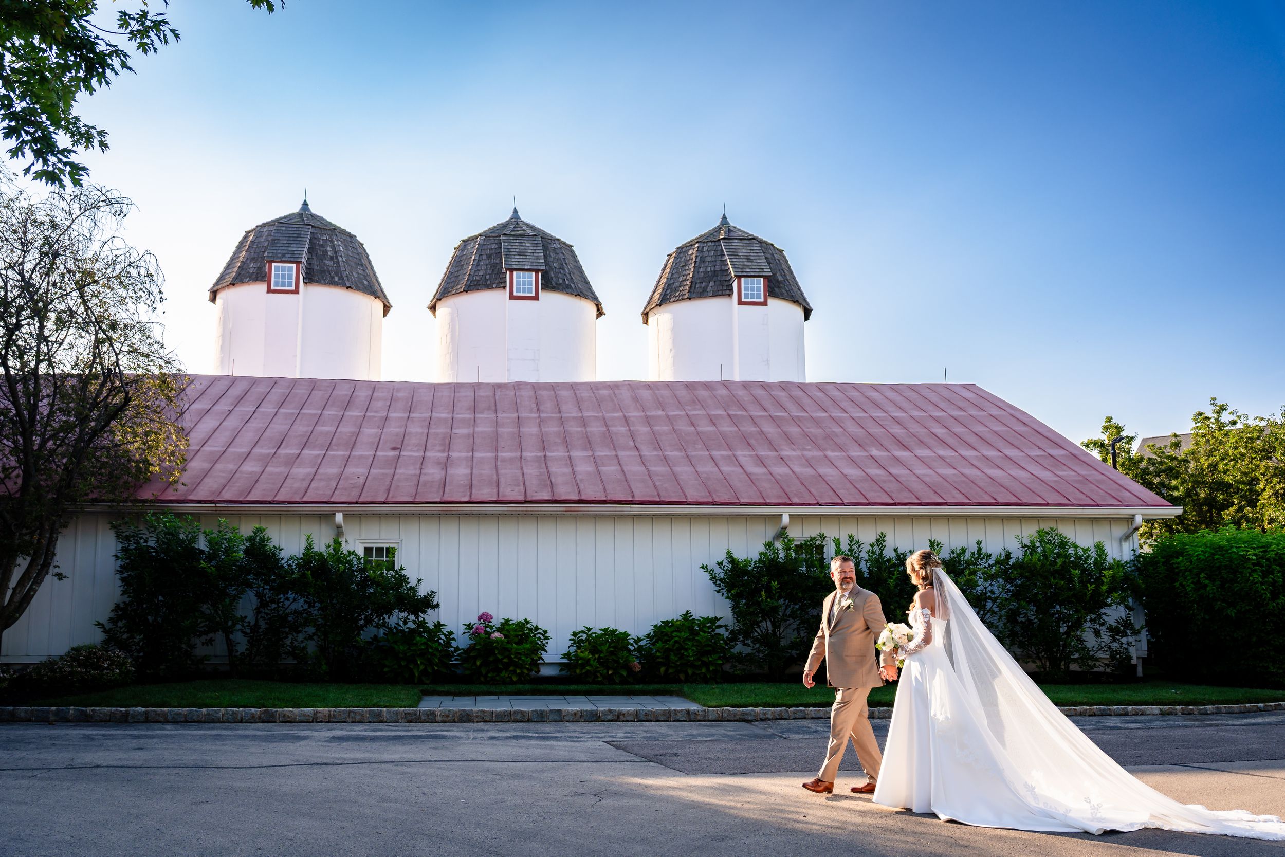 Bride and groom walk hand in hand toward a white barn wedding venue under a clear blue sky.
