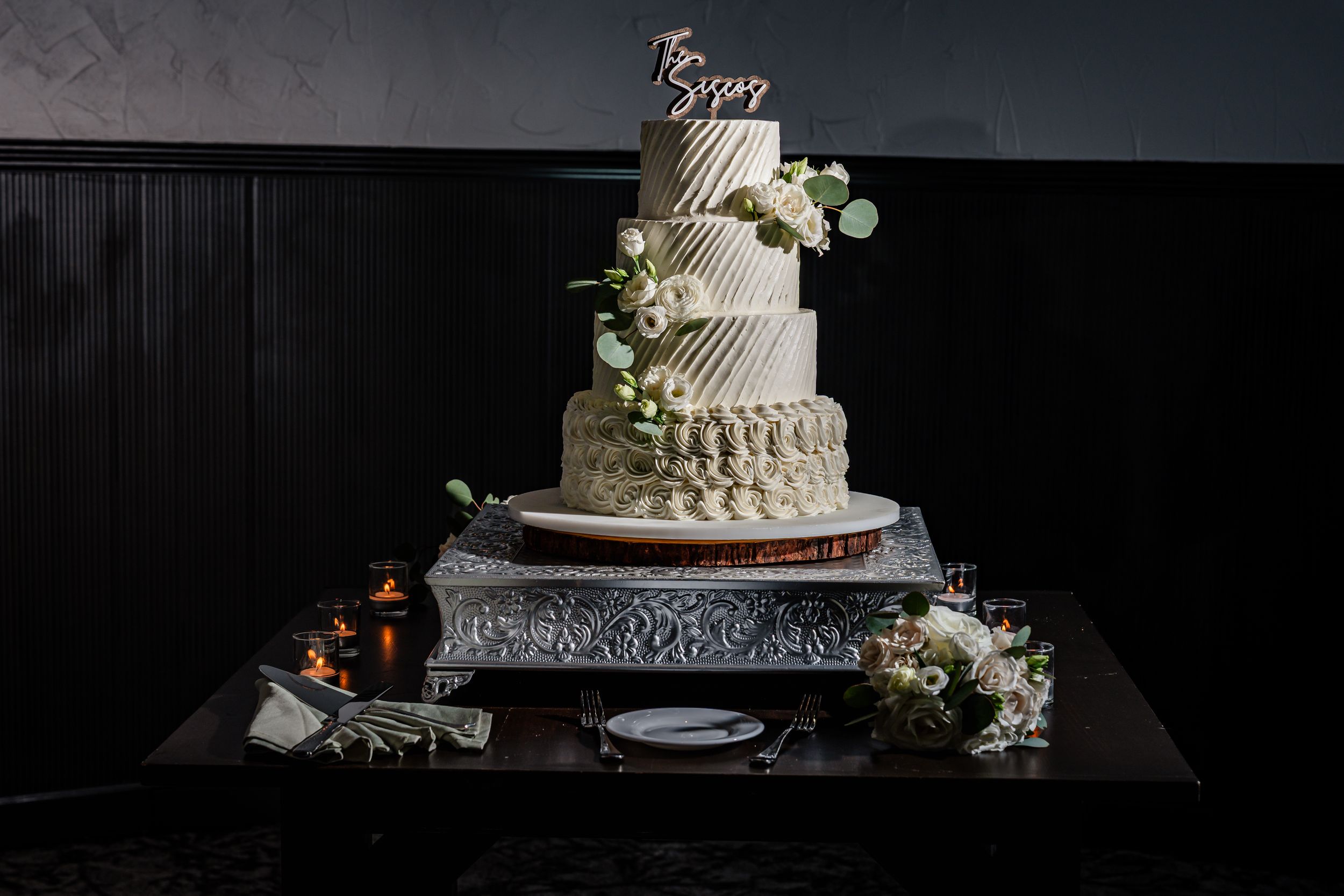 Elegant four-tier white wedding cake adorned with greenery and floral topper displayed on silver stand.