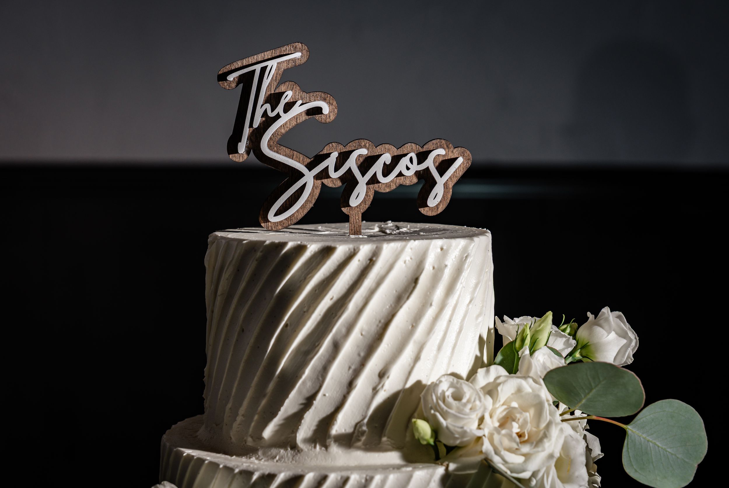 Elegant three-tier white wedding cake with textured frosting, white roses, and a rose gold 'The Siscos' cake topper.