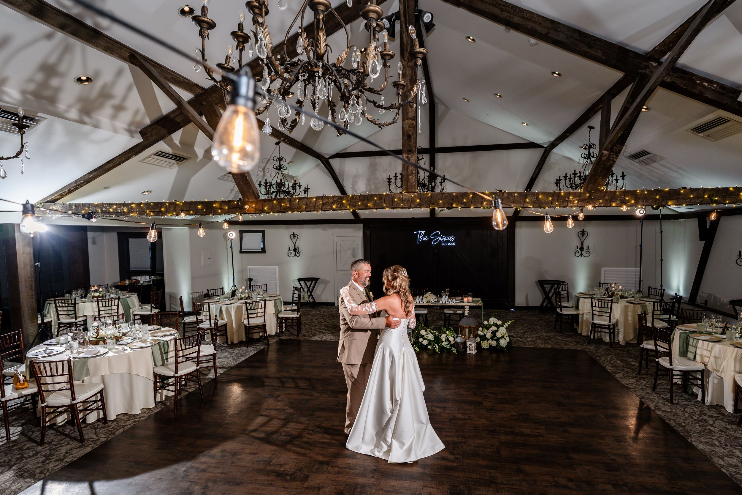 Bride and groom share first dance in elegant rustic wedding venue with chandeliers, string lights, and decorated reception tables.