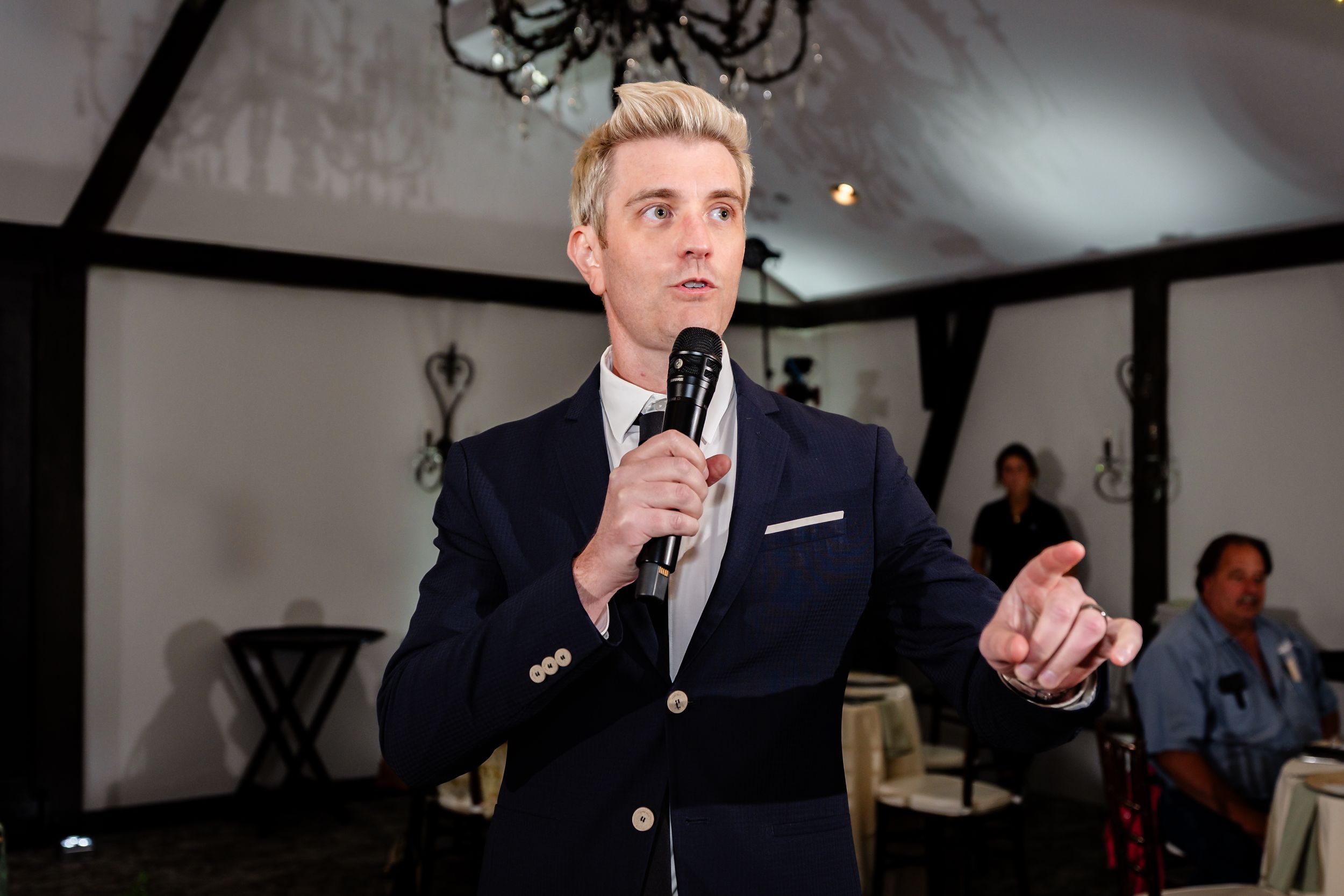 Man in navy blazer speaking into microphone at elegant indoor event.