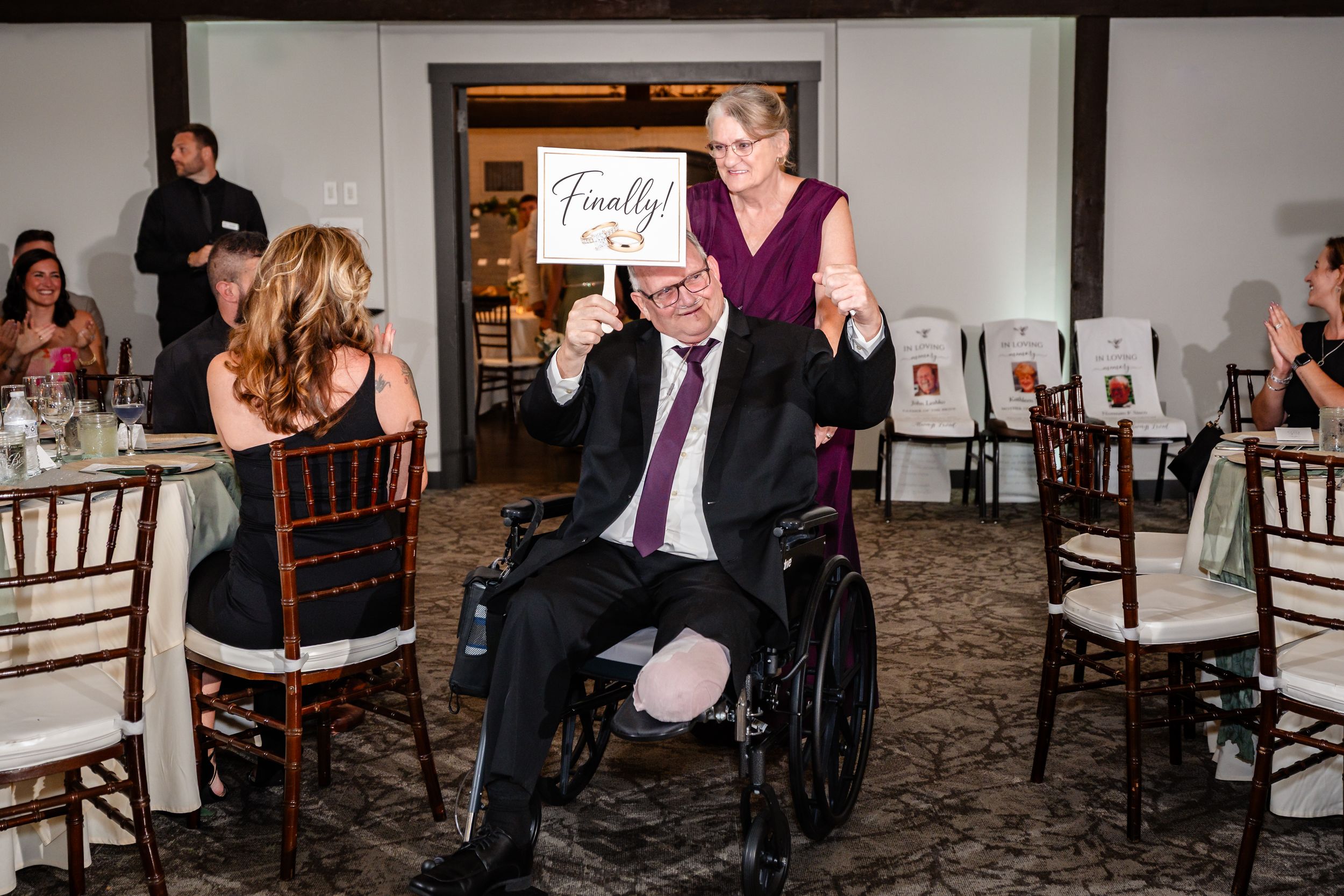 Man in wheelchair gives thumbs up at wedding reception holding Finally sign while woman stands behind him smiling.