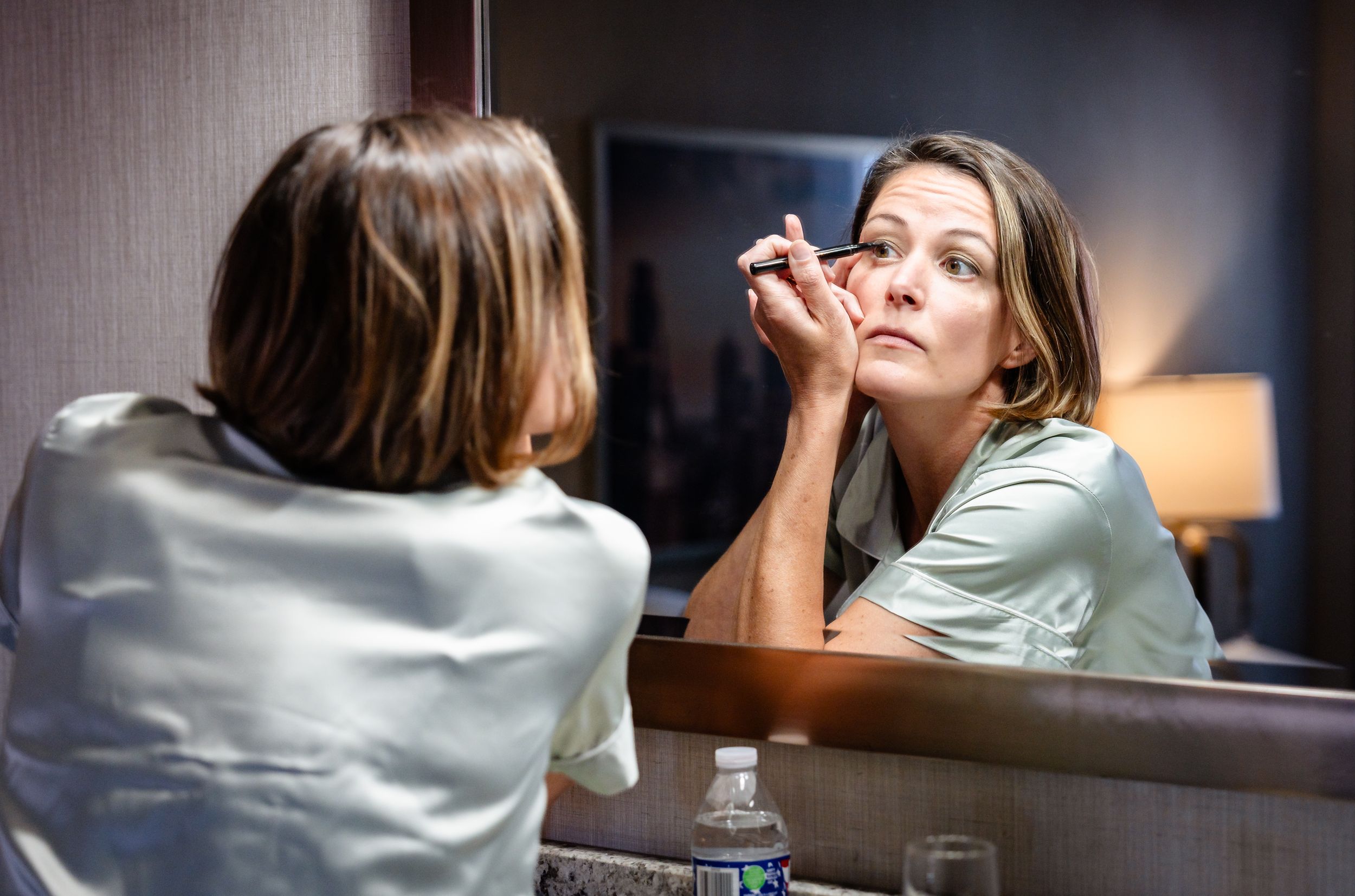 Woman applies eye makeup while looking into a hotel mirror during wedding day preparations.
