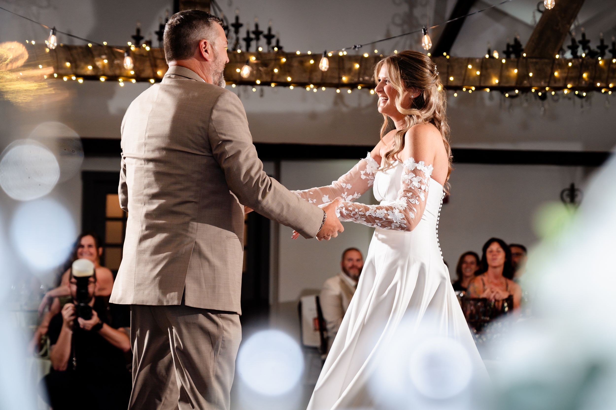 Bride and groom share first dance under warm string lights surrounded by wedding guests.