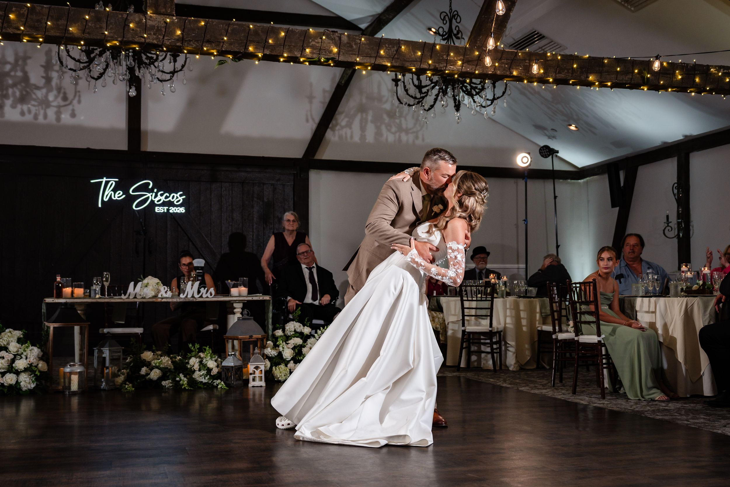 Bride and groom share romantic first dance dip in elegant wedding reception venue with string lights and chandelier.