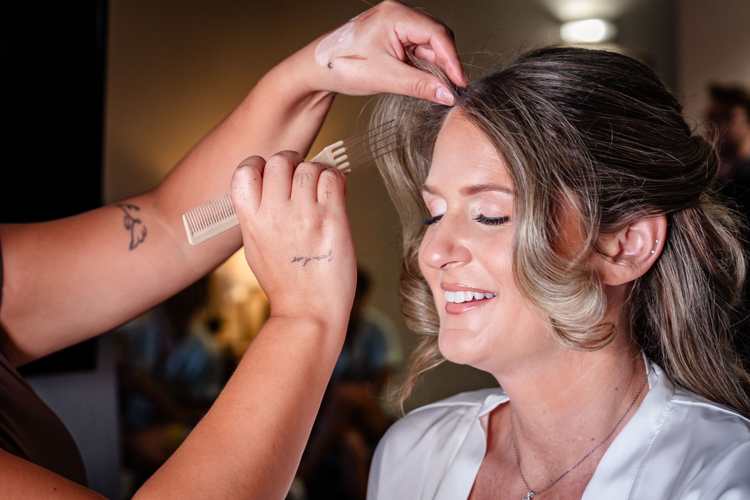 Bride smiling as hairstylist styles her hair during wedding day preparations in a bright dressing room.