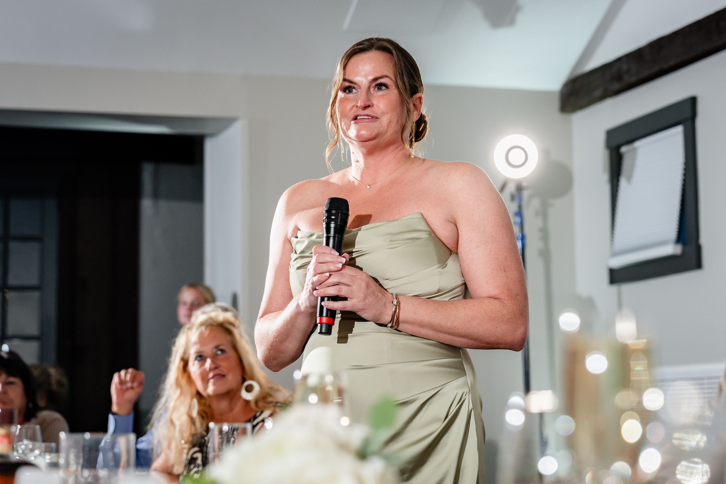 Bridesmaid in sage green strapless gown delivers a heartfelt speech at an elegant wedding reception.