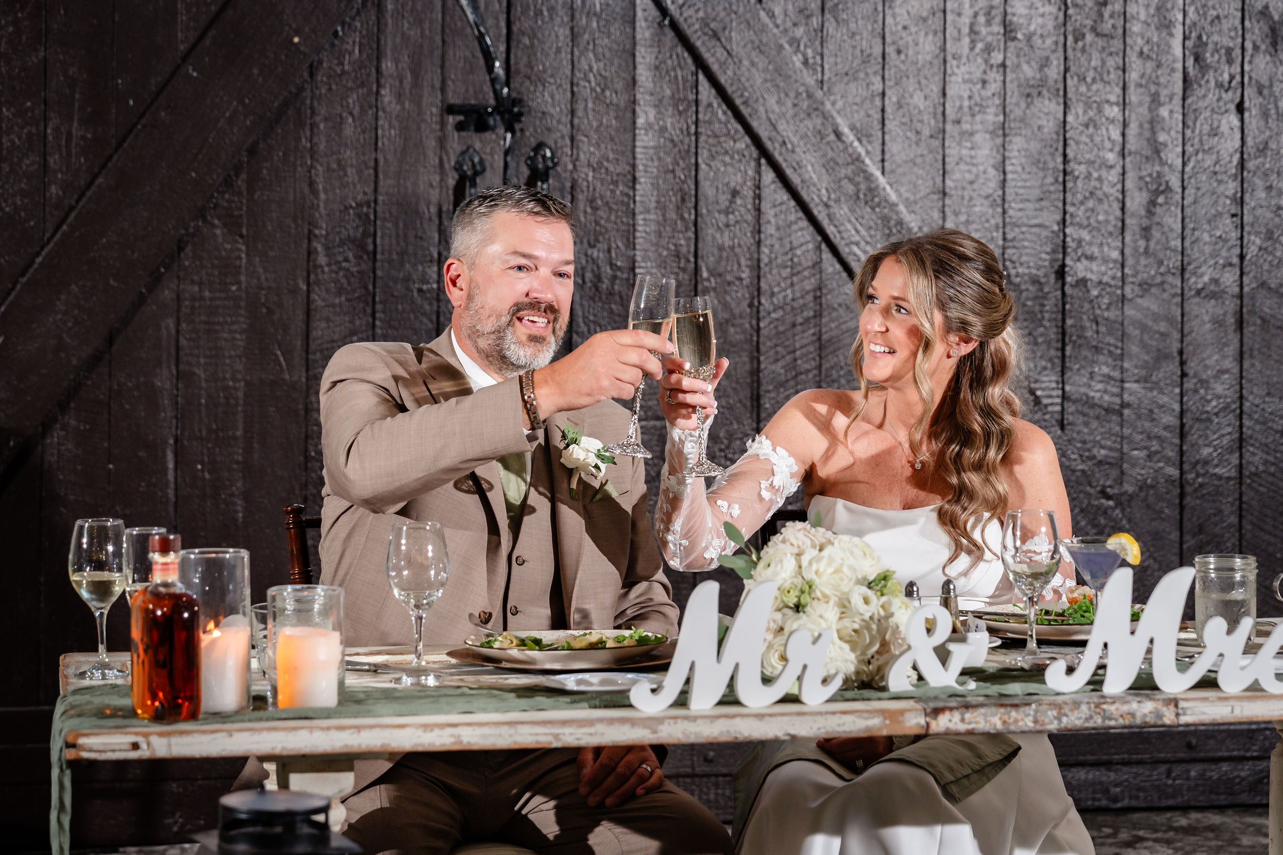 Bride and groom toast champagne glasses at rustic barn wedding reception table with Mr & Mrs sign.