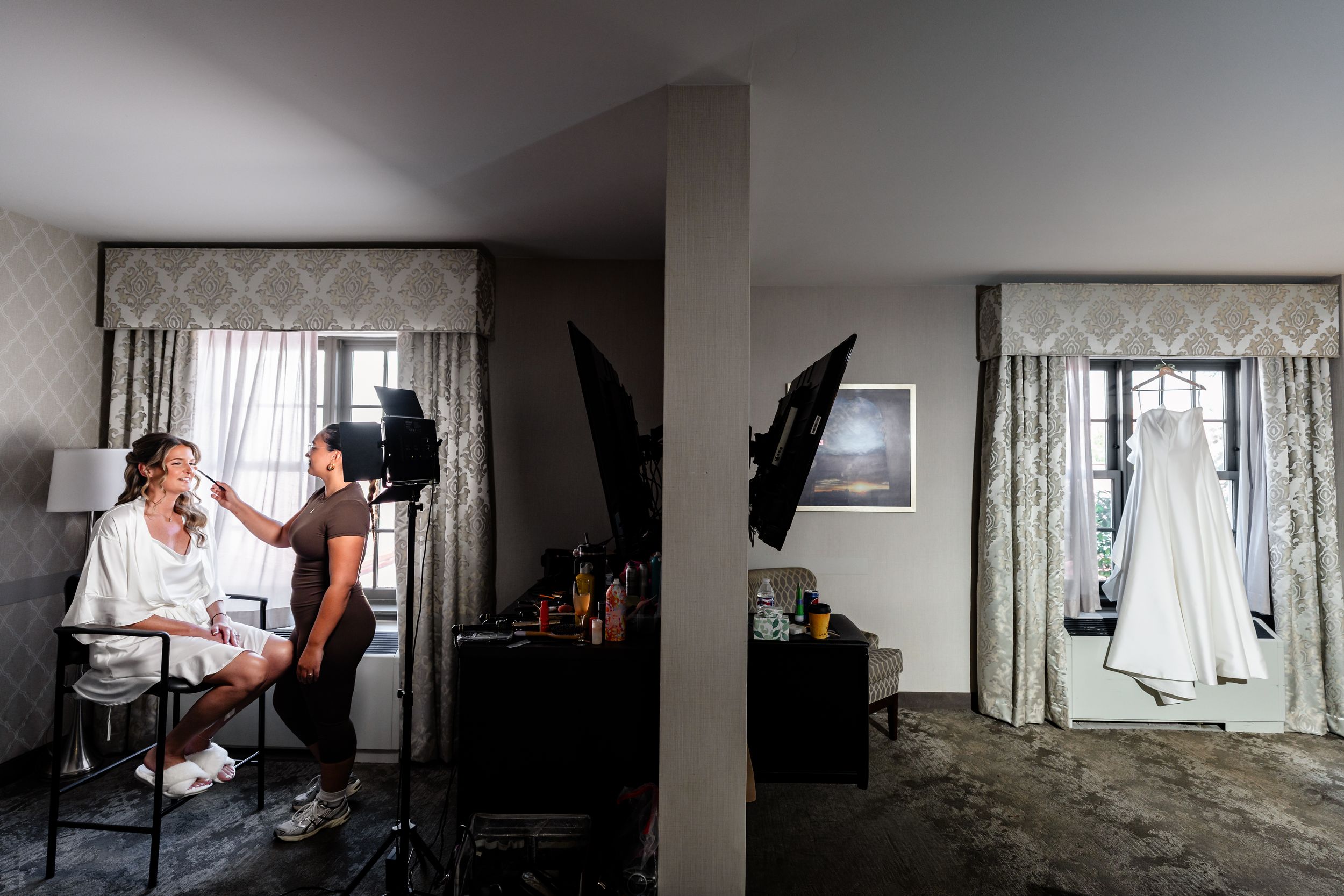 Bride getting makeup done in hotel room with wedding dress hanging in background.