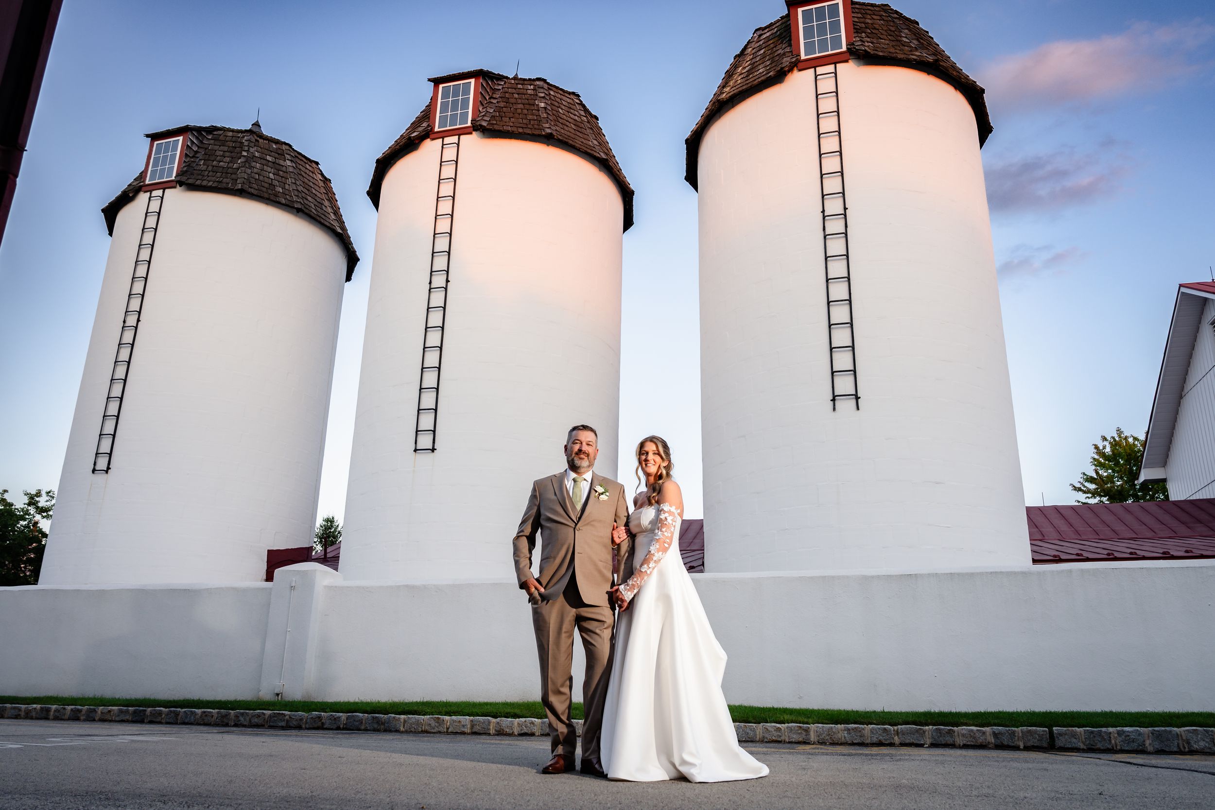 Wedding couple poses in front of white grain silos at golden hour outdoor venue.
