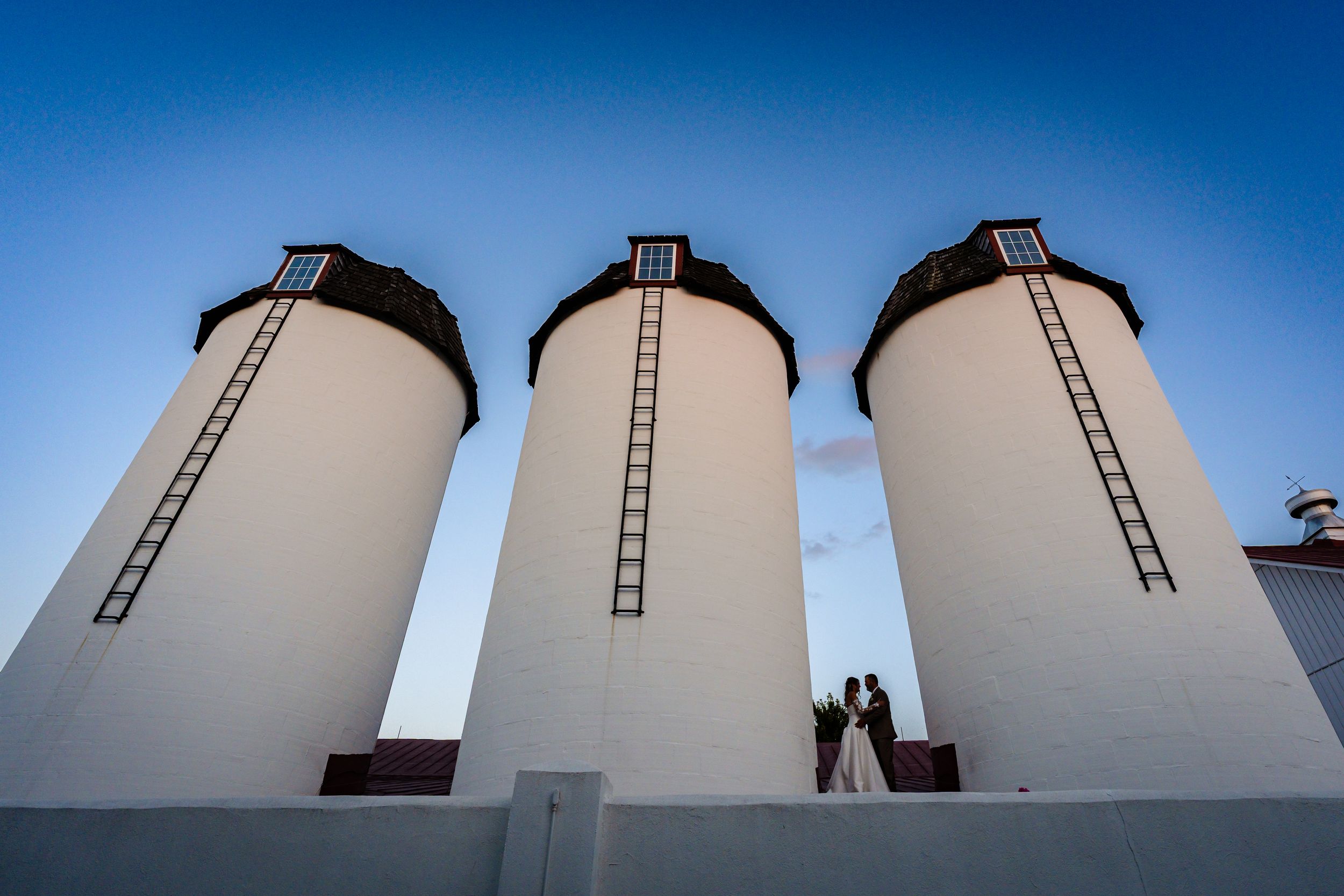 Bride and groom silhouetted against blue sky beside three tall white grain silos with ladders.
