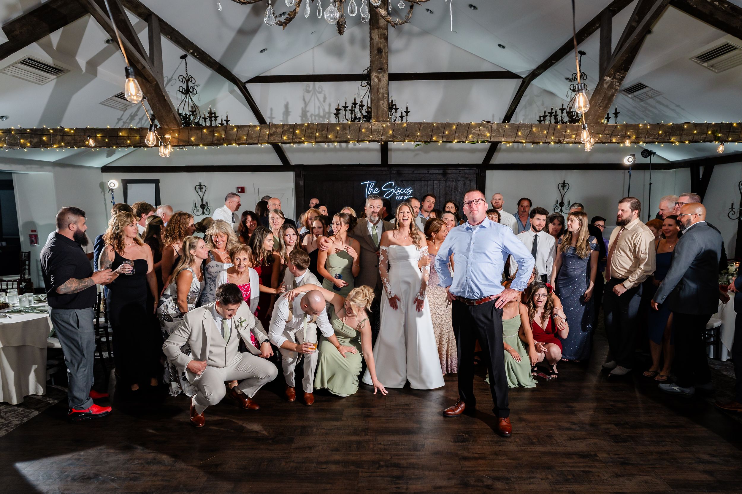 Wedding reception group photo with bride and groom posing with guests in an elegant rustic venue with string lights.