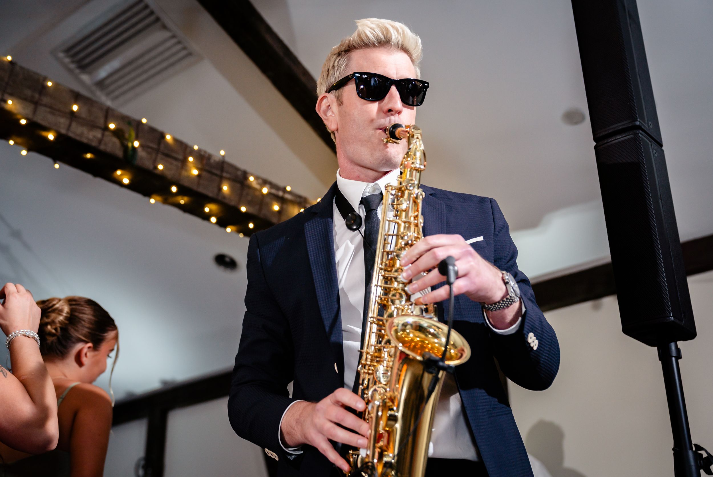 Blonde saxophonist in tuxedo and sunglasses performing at elegant wedding reception with string lights.