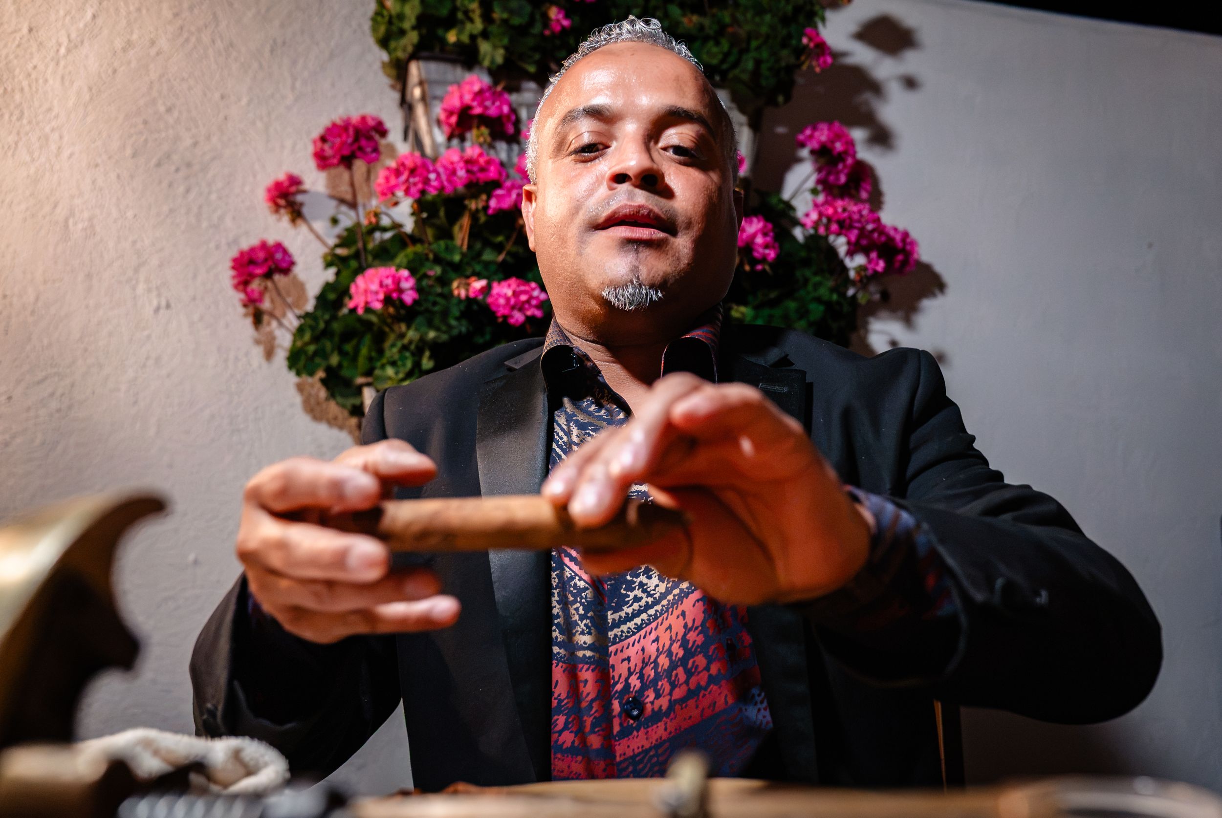 Man in blazer holding a cigar at a table with pink flowers in the background.