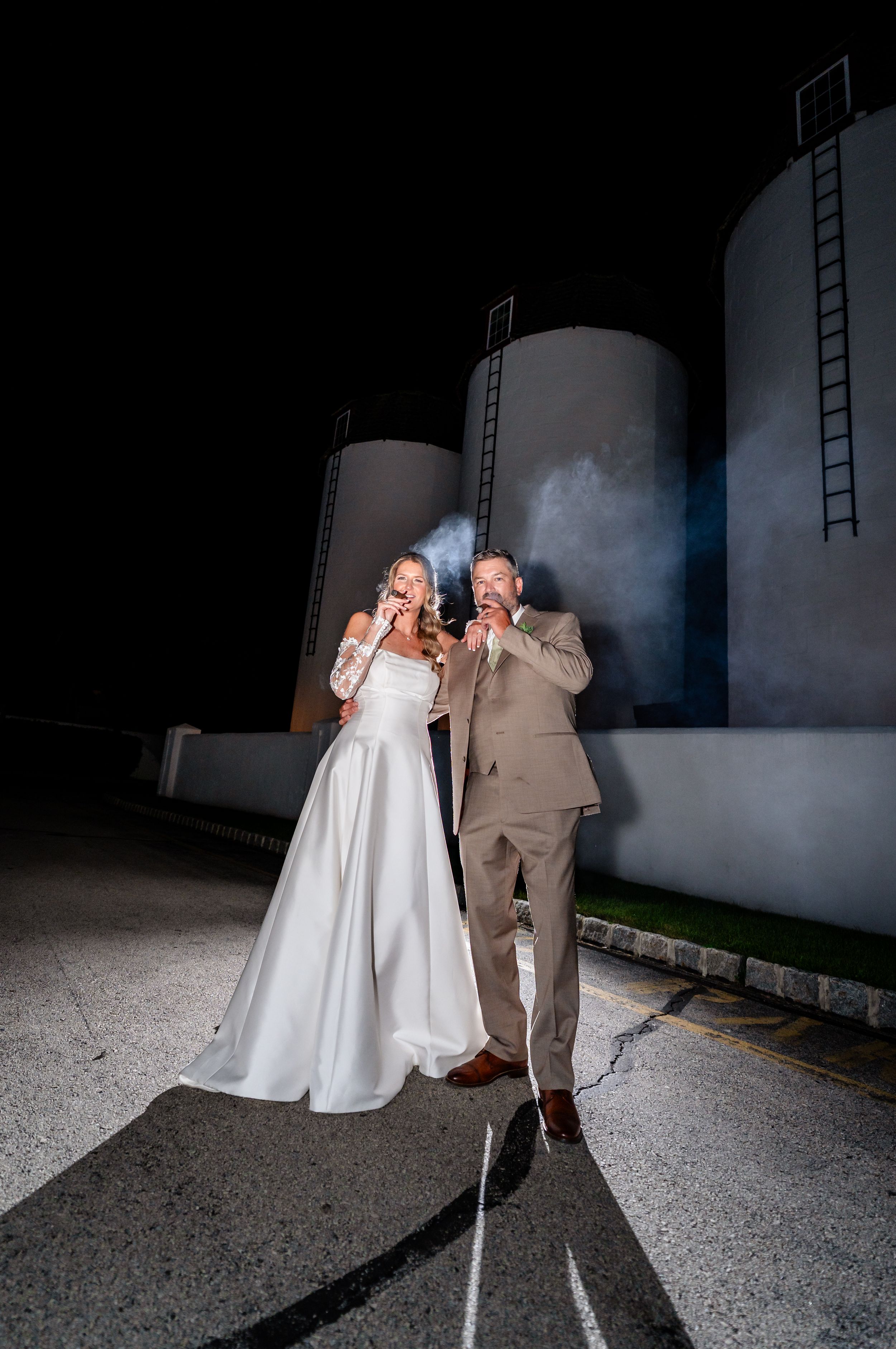 Bride and groom smoking cigars at night near industrial silos in wedding attire.