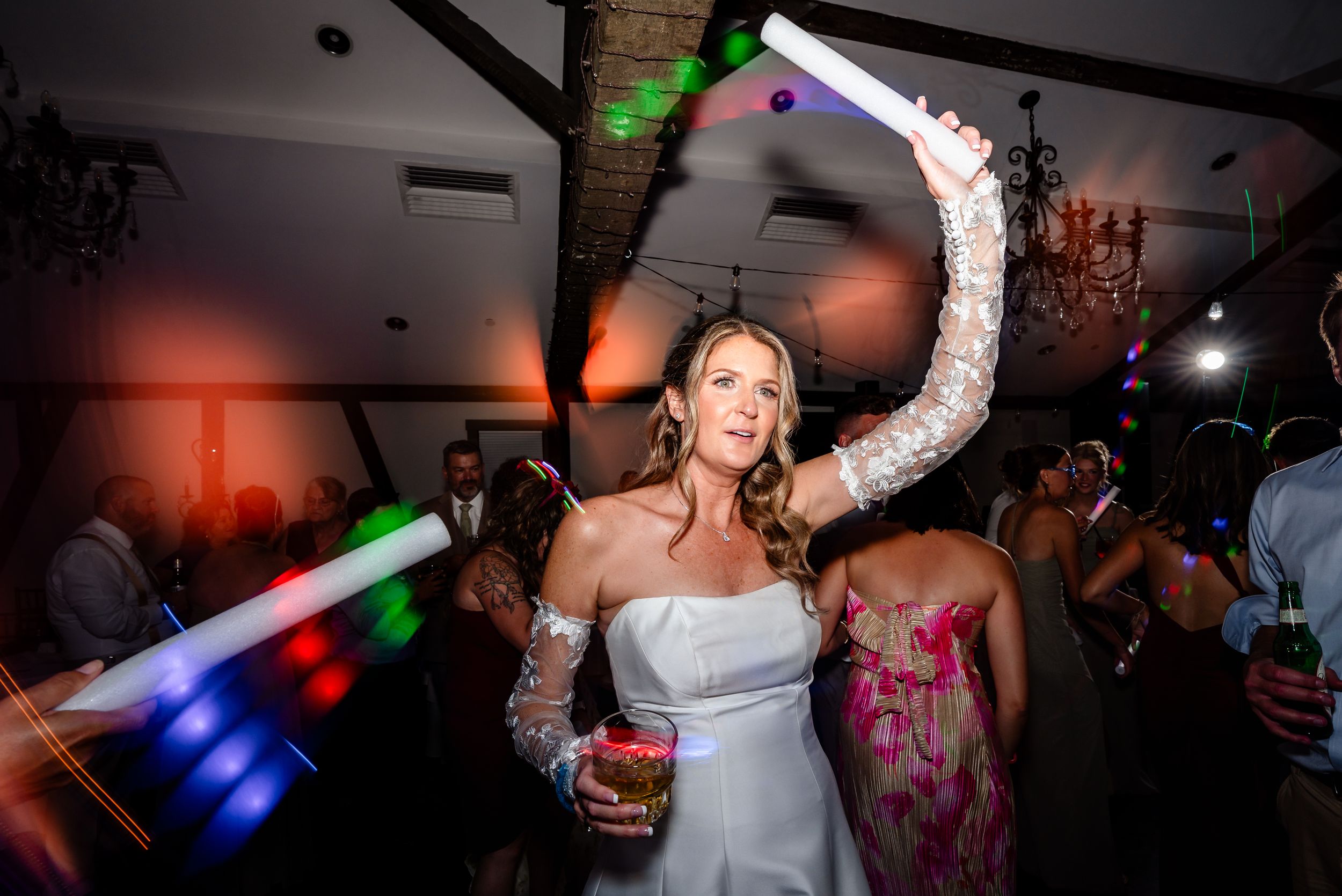 Bride dancing joyfully at wedding reception holding a light-up foam stick while guests celebrate around her.