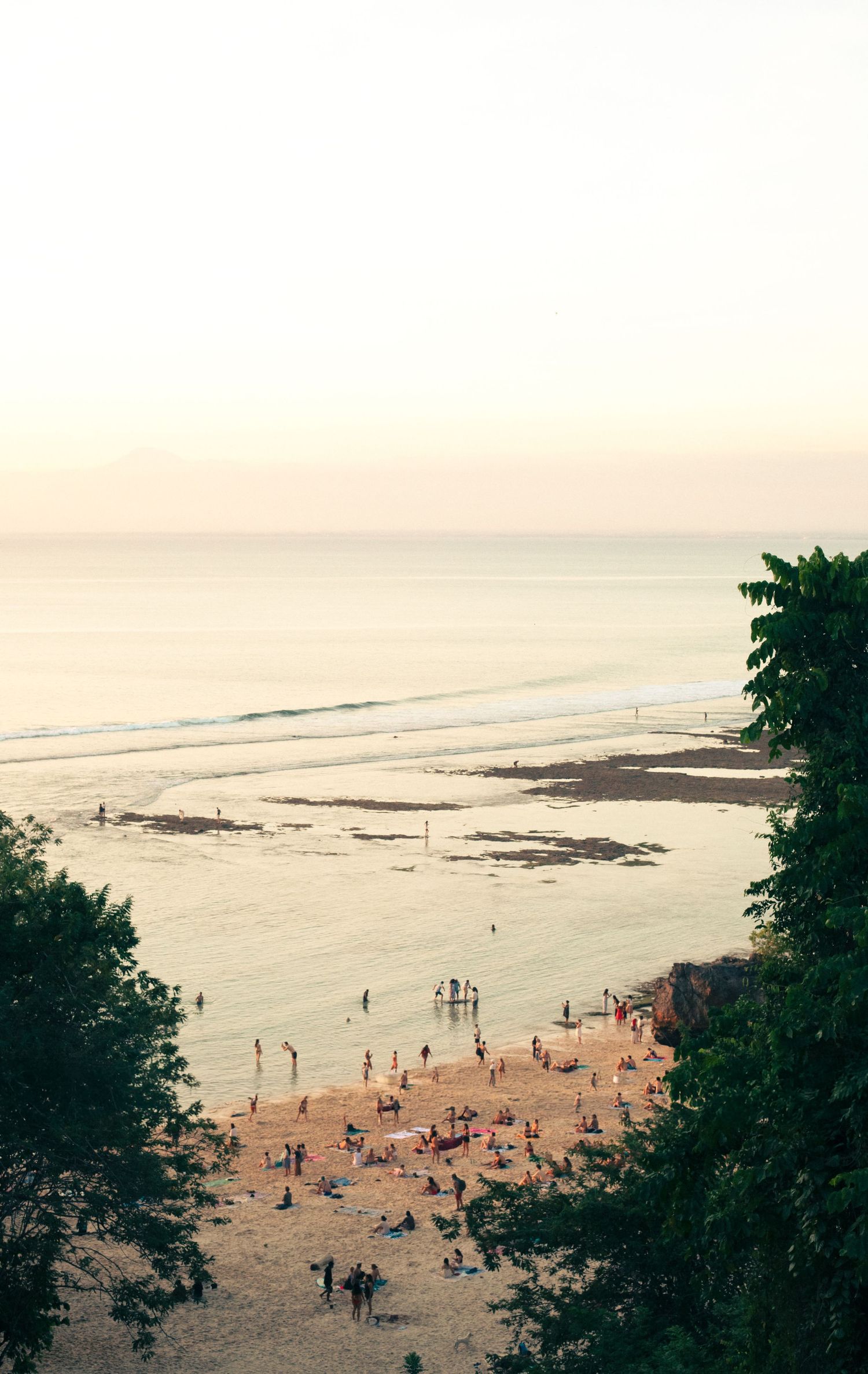 Aerial view of a crowded tropical beach at sunset with calm ocean waves and lush green trees framing the shore.