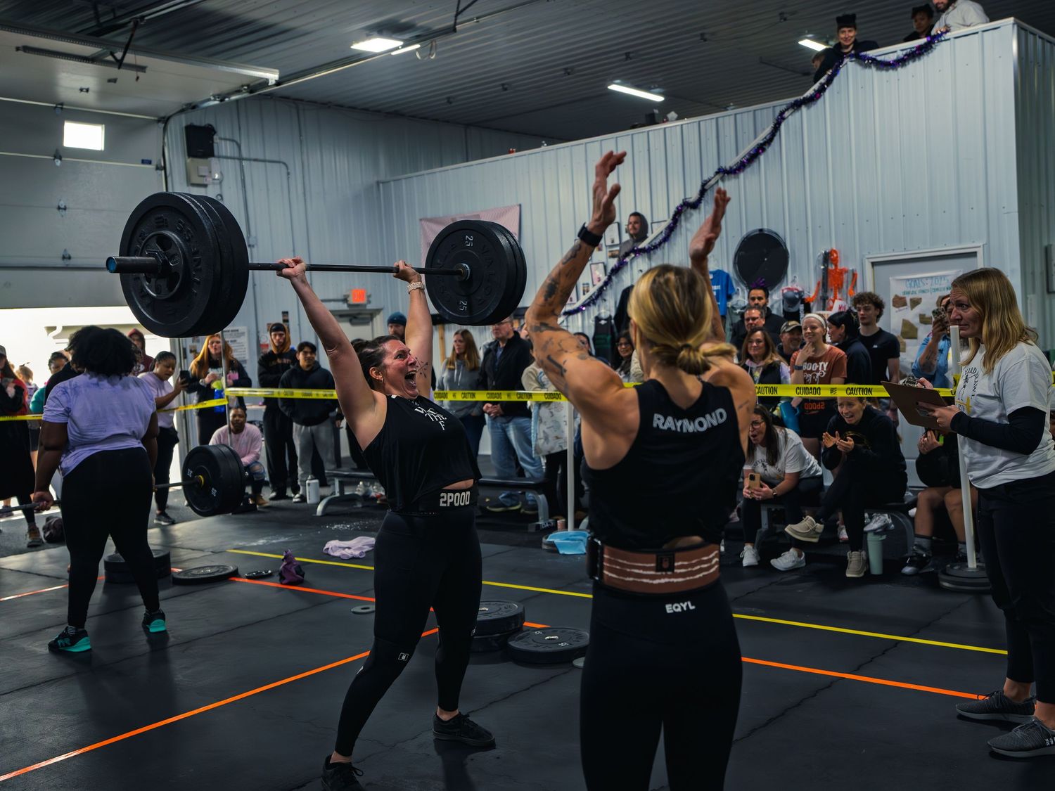 CrossFit athlete grinds a loaded barbell to lockout overhead, face straining, as a judge counts reps and the crowd watches at a competition.