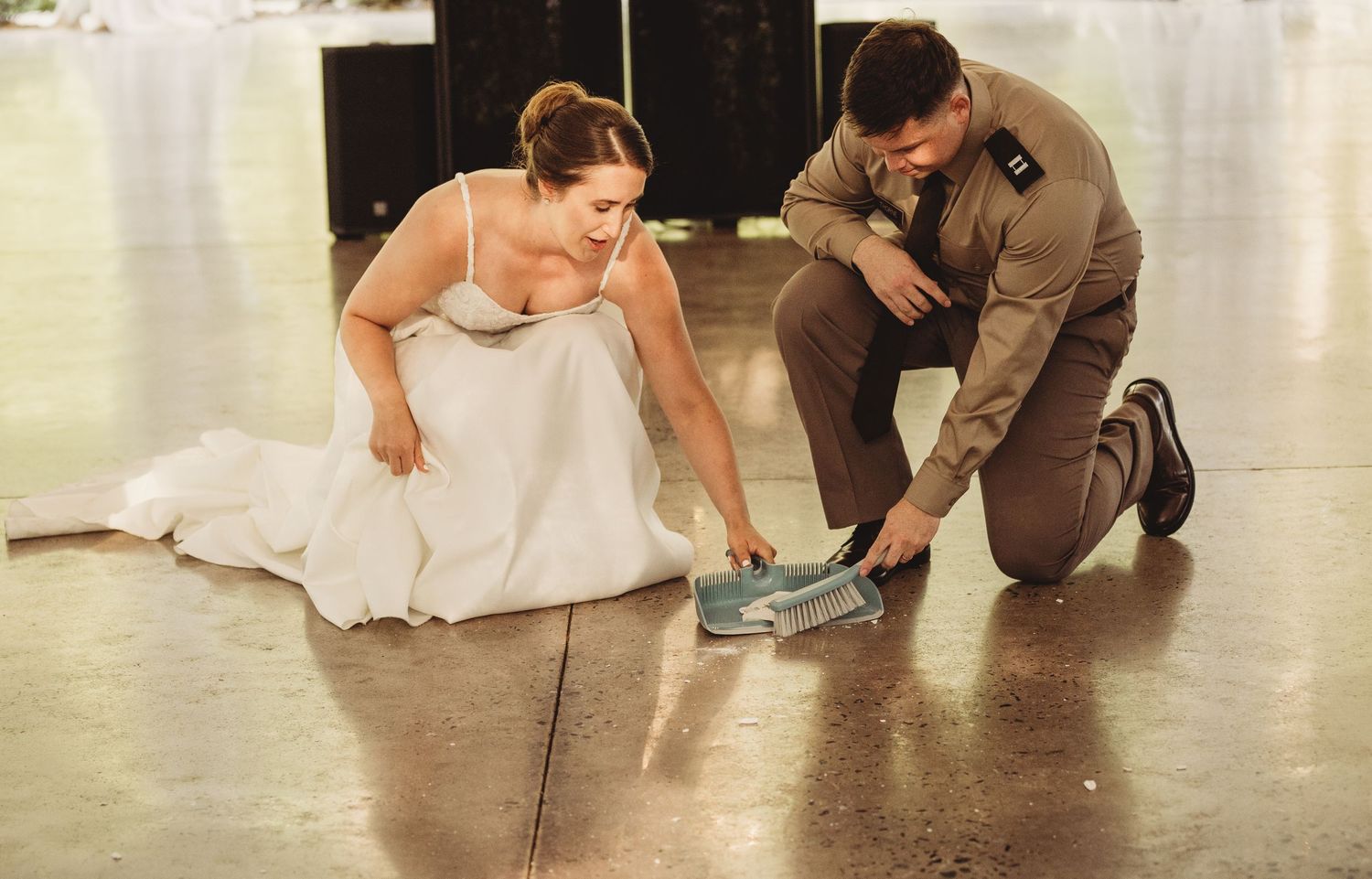 Bride in white gown and groom in military uniform kneel on dance floor examining something together.