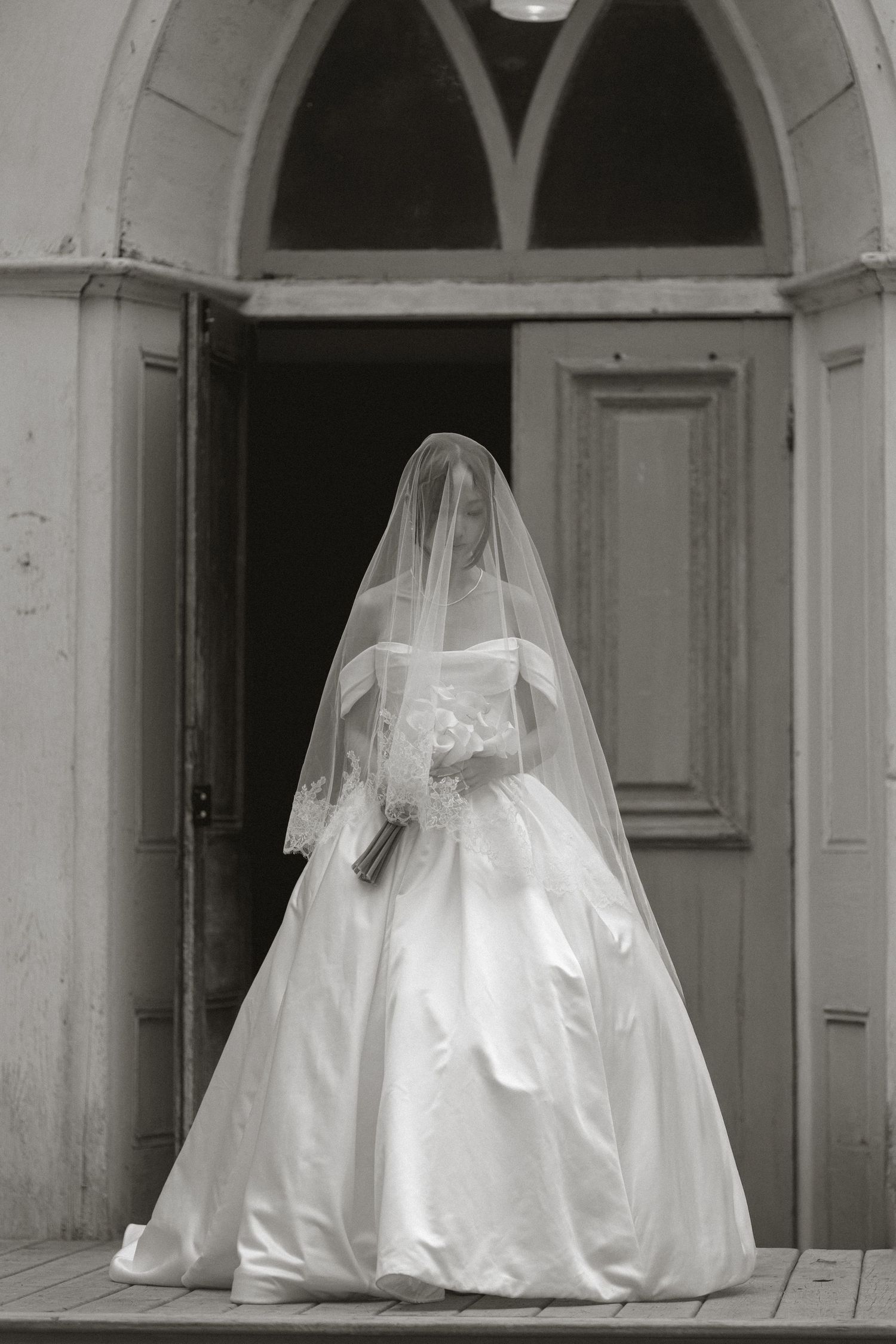 Bride in a full white ball gown and veil standing at church doorway in black and white photo.