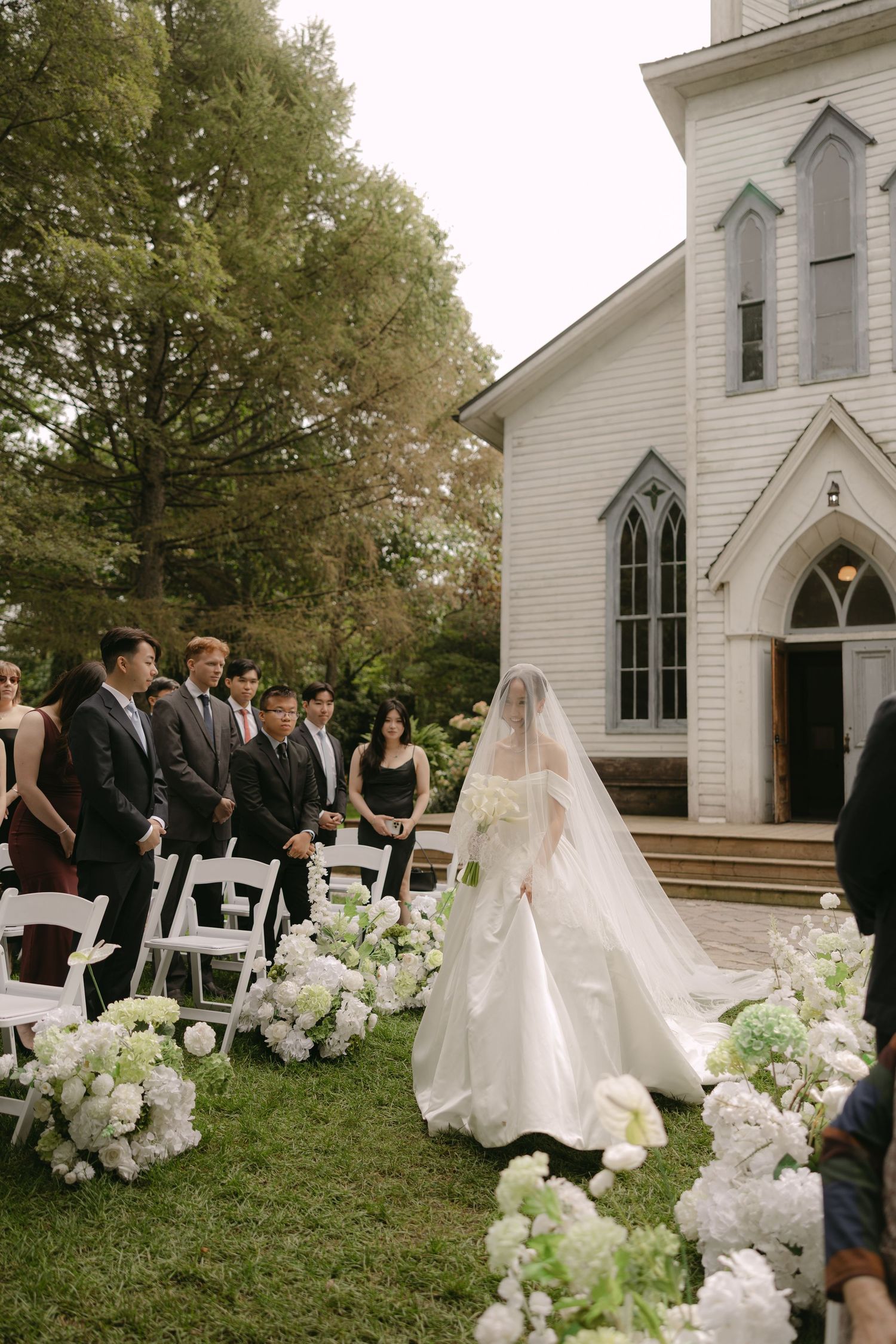 Bride in white gown walks down floral aisle toward white chapel during outdoor wedding ceremony.