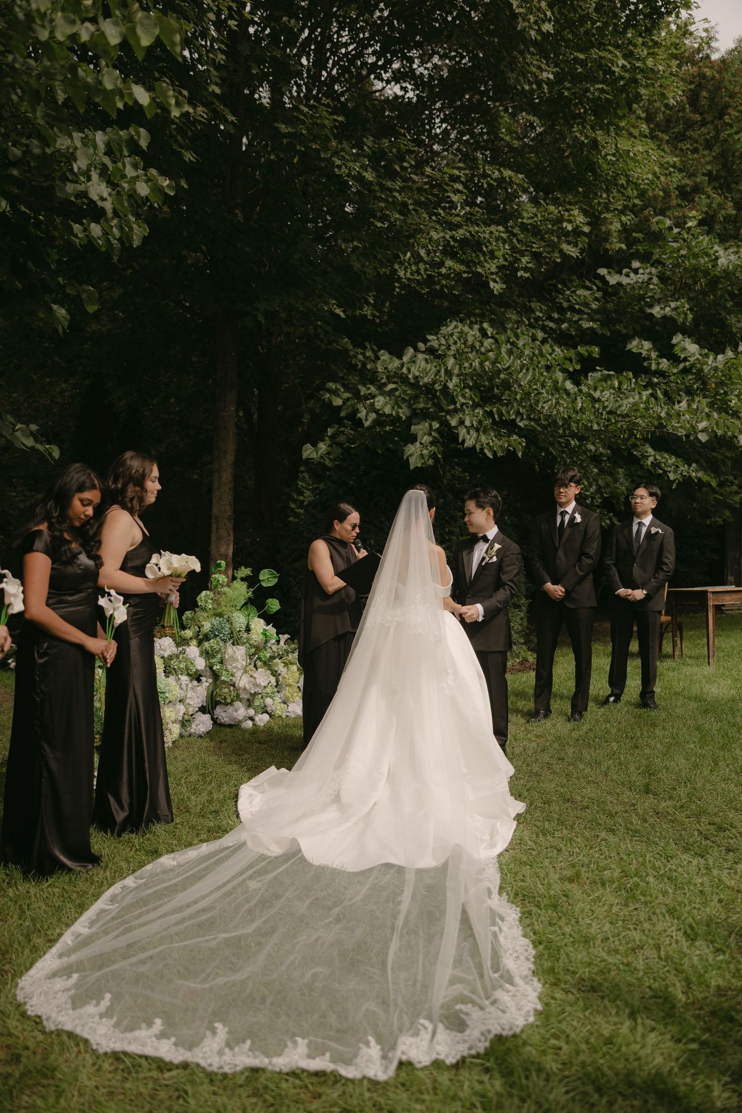 Bride in cathedral veil faces groom at outdoor garden ceremony with bridesmaids in black gowns.