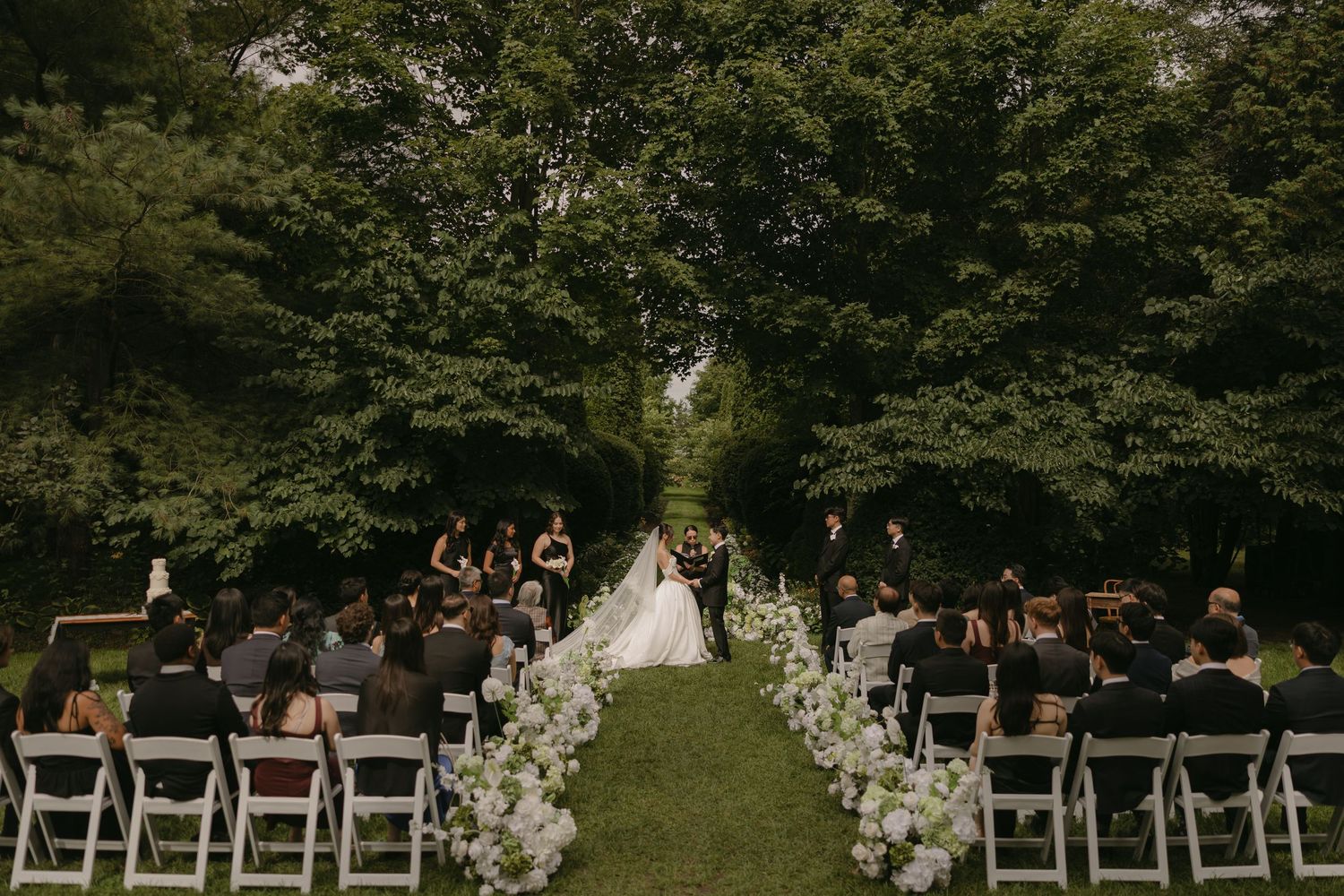 Aerial view of elegant outdoor wedding ceremony on green lawn lined with white floral arrangements.