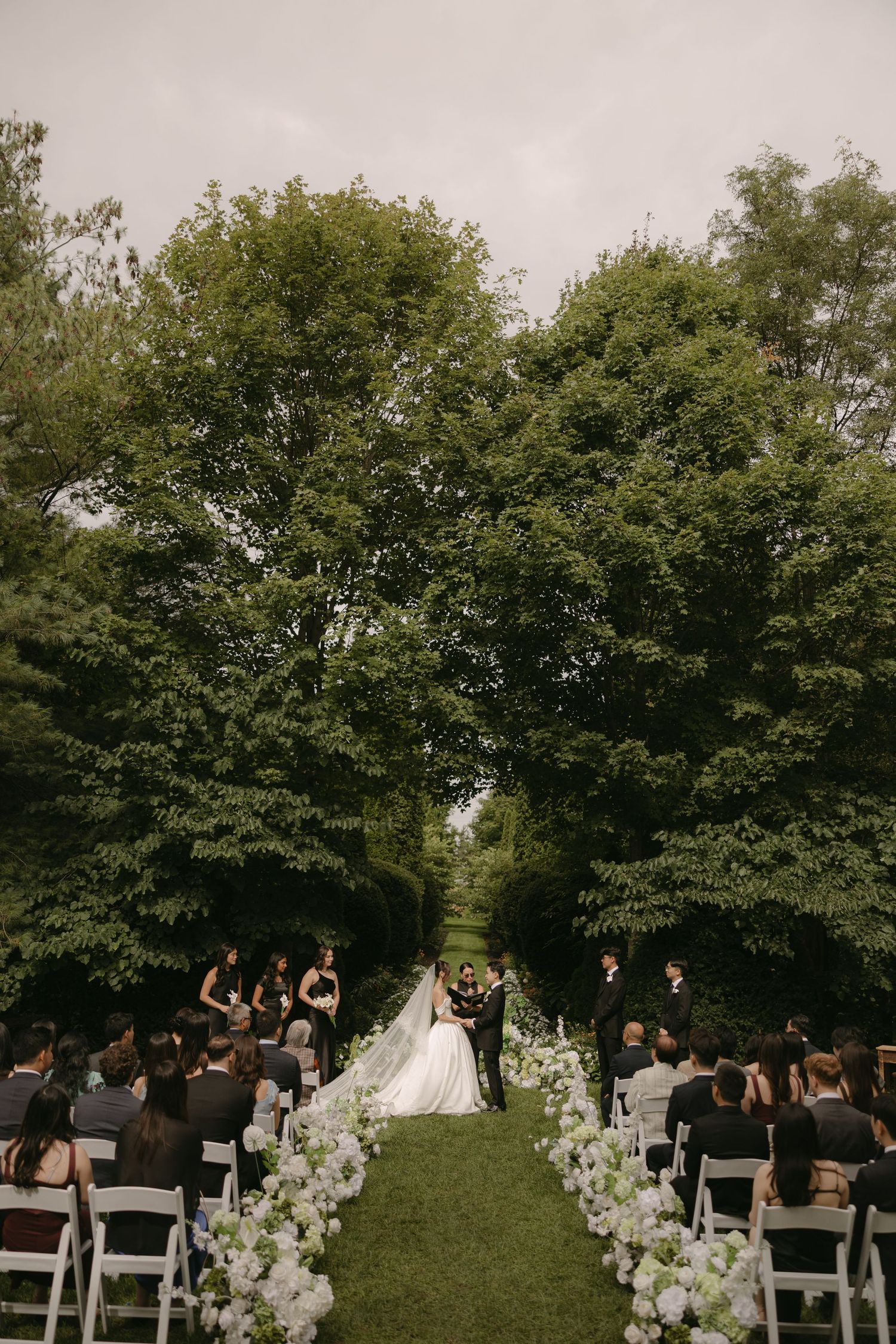 Wide shot of outdoor wedding ceremony flanked by lush trees and white hydrangea floral aisle decor.