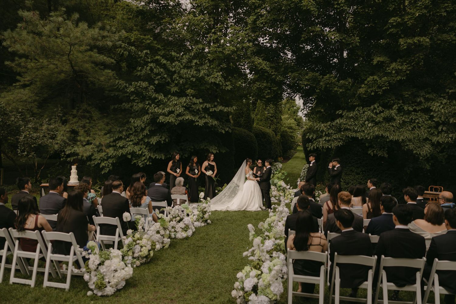 Couple exchanges vows at outdoor garden wedding surrounded by guests and white floral arrangements.