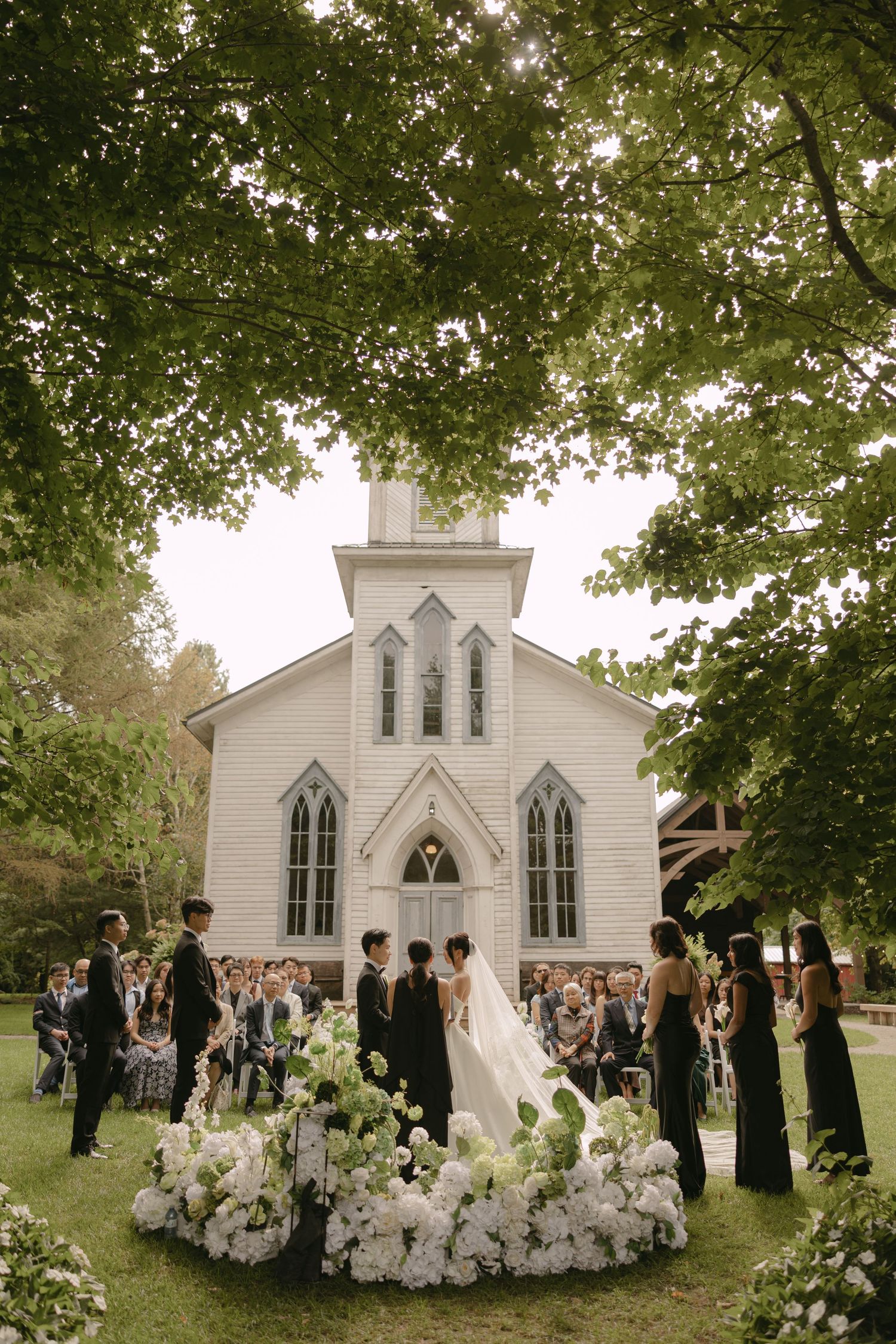 Bride and groom stand before white chapel surrounded by guests and white floral circle arrangement.