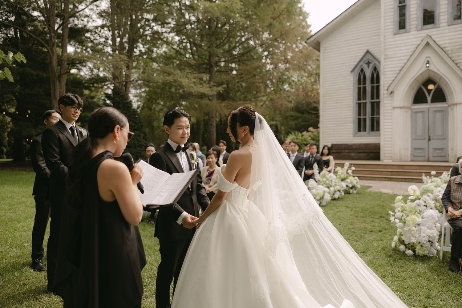 Groom reads vows to bride in cathedral veil outside white chapel during garden wedding ceremony.