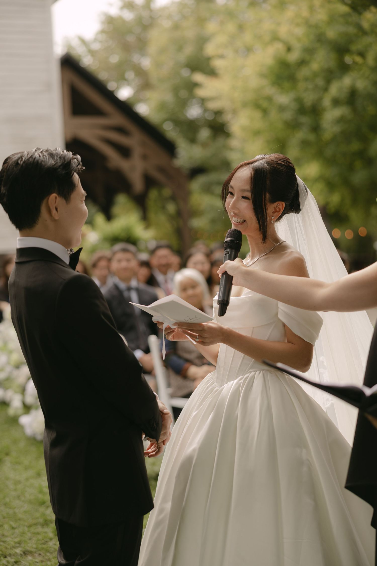 Bride reads wedding vows from paper while holding microphone at outdoor garden ceremony.