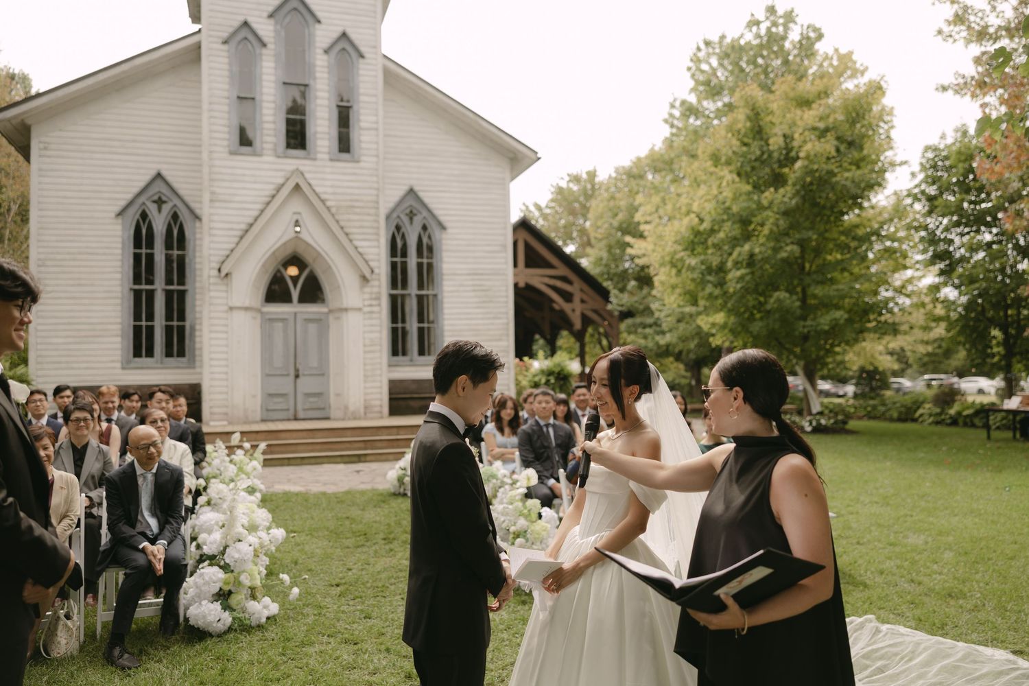 Couple exchanges vows outdoors with white chapel and lush trees in background.