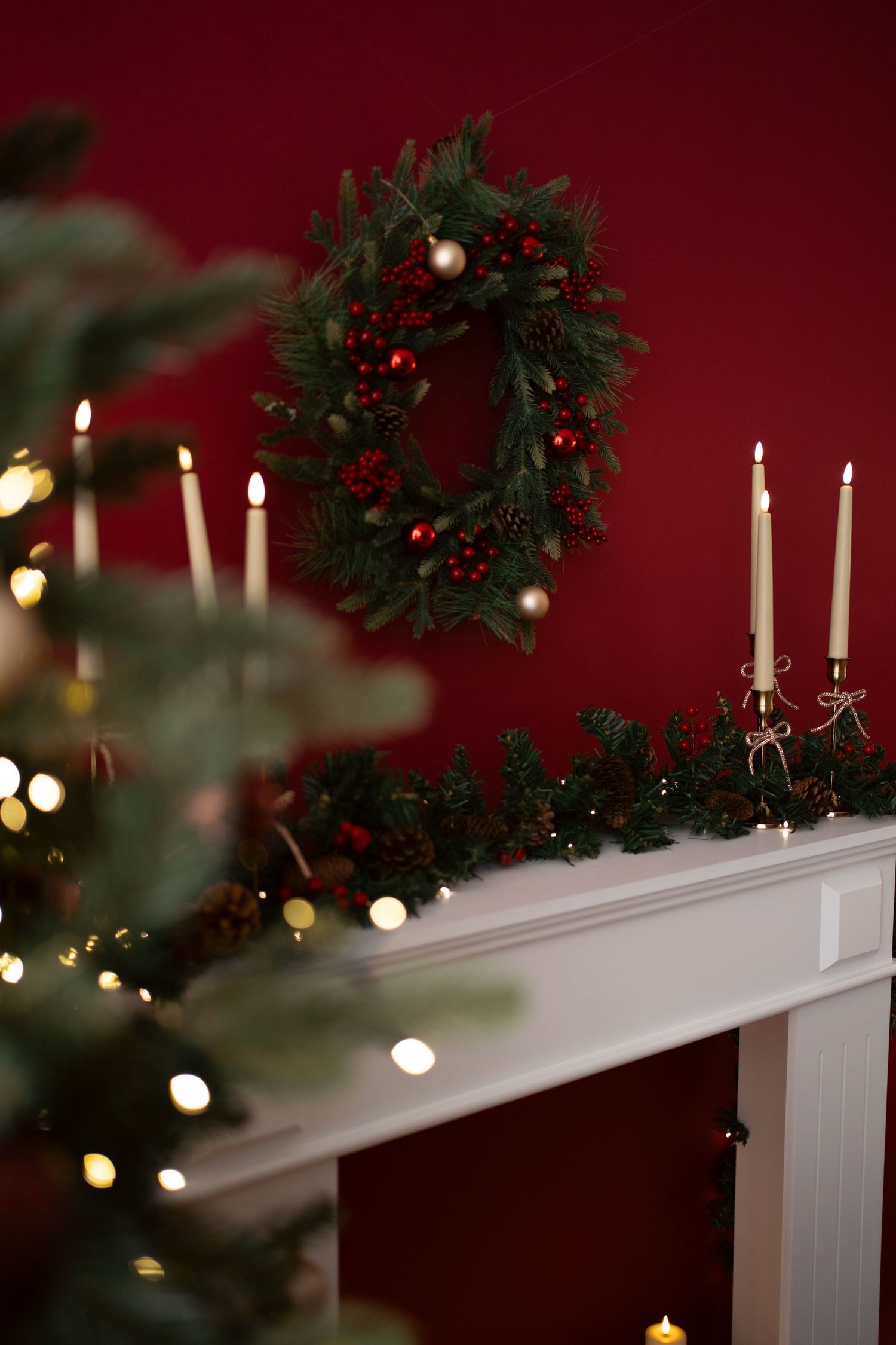 Christmas wreath with red berries and ornaments on a red wall above a white fireplace mantel with candles and garland.