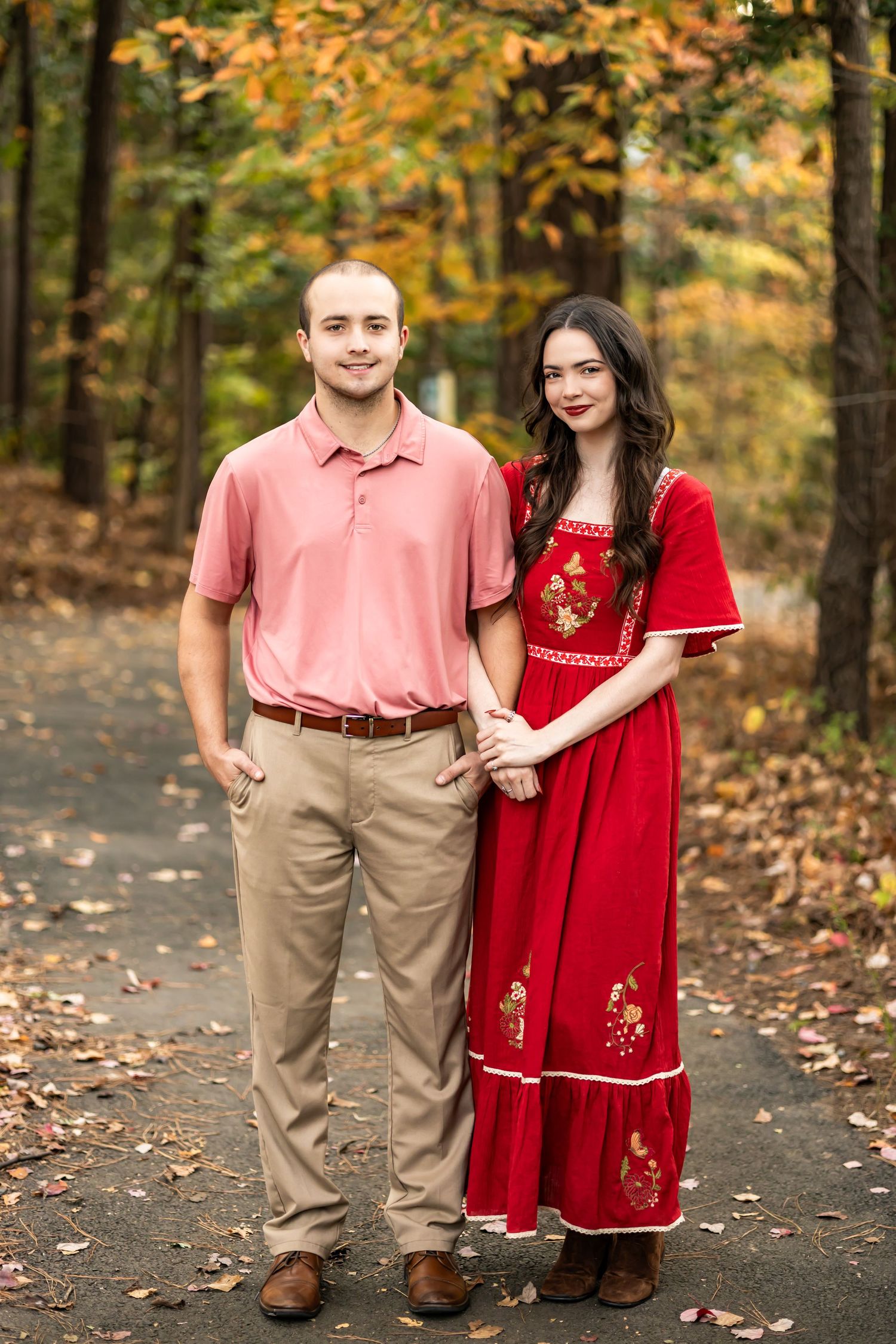 Couple posing on autumn path, man in pink polo and khakis, woman in red embroidered dress, fall foliage background.