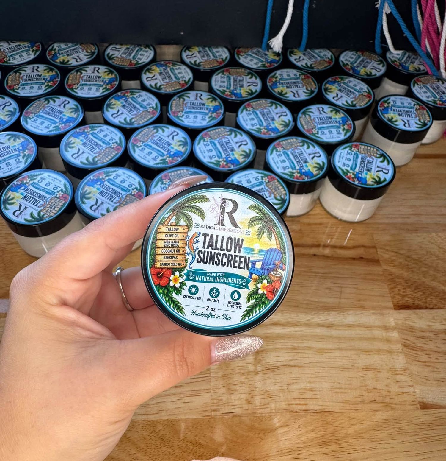 Hand holding a jar of tallow sunscreen with tropical label, surrounded by multiple product jars on a table.