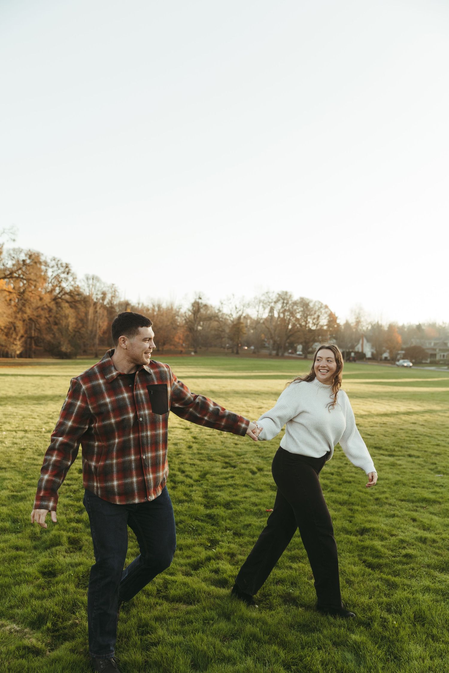 A couple shares a tender kiss outdoors at golden hour, woman in white sweater embracing man in plaid jacket.