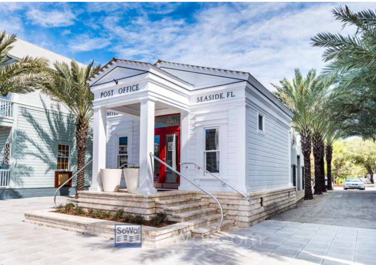 White post office building in Seaside, FL, featuring a red door, palm trees, and classic Southern architecture under blue skies.